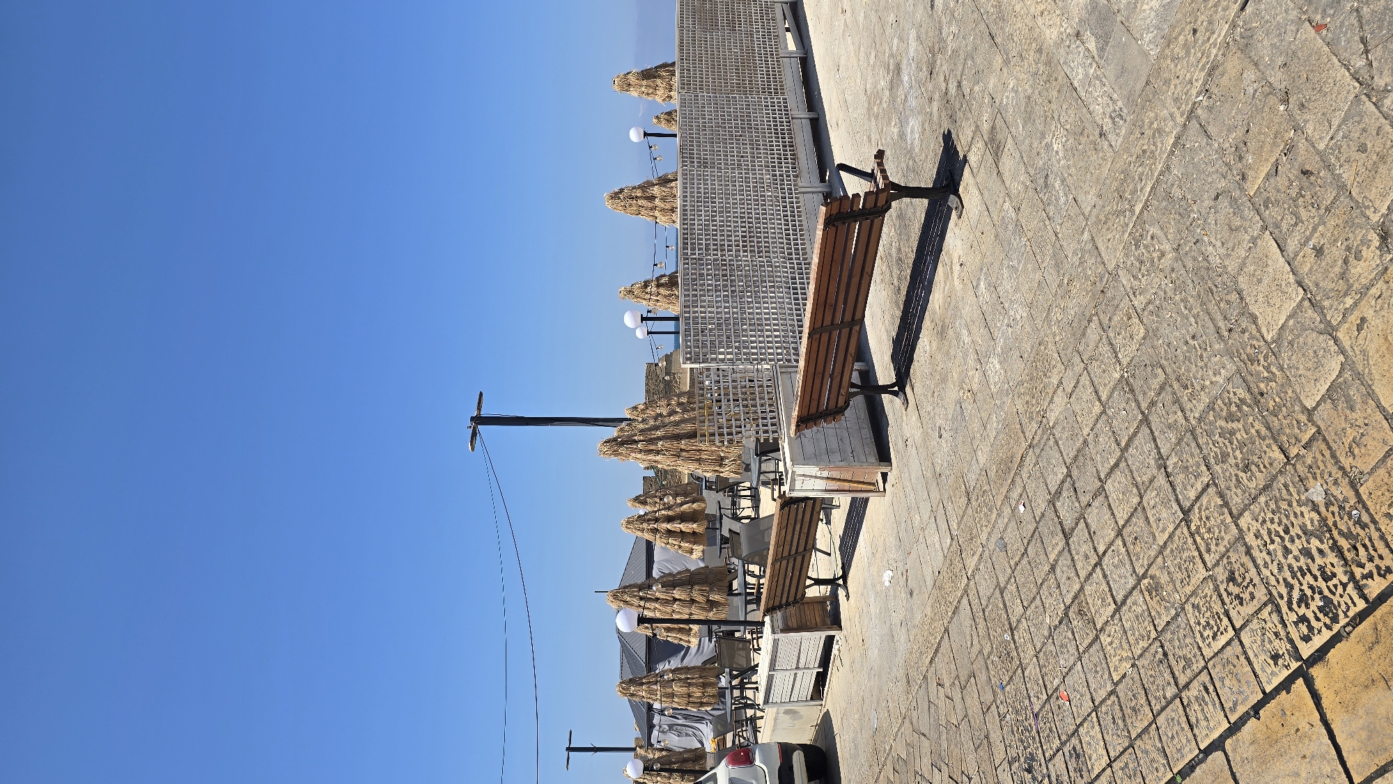 This image depicts an outdoor seating area at a coastal location, likely a cafe or restaurant, in the city of Acre, Israel. The foreground is paved with large, worn stone tiles. Two empty wooden benches are positioned on the paving, with a long shadow cast by the bench in the mid-ground. Behind the benches, a white lattice fence divides the seating area from the rest of the space. Several tall, straw-covered umbrellas are spaced out, resembling pine trees. Tables and chairs are visible under these umbrellas, some covered by a gray tarp. A white car is parked to the left of the frame. The background shows a clear blue sky above the sea. The scene appears to be unoccupied, with no people present.