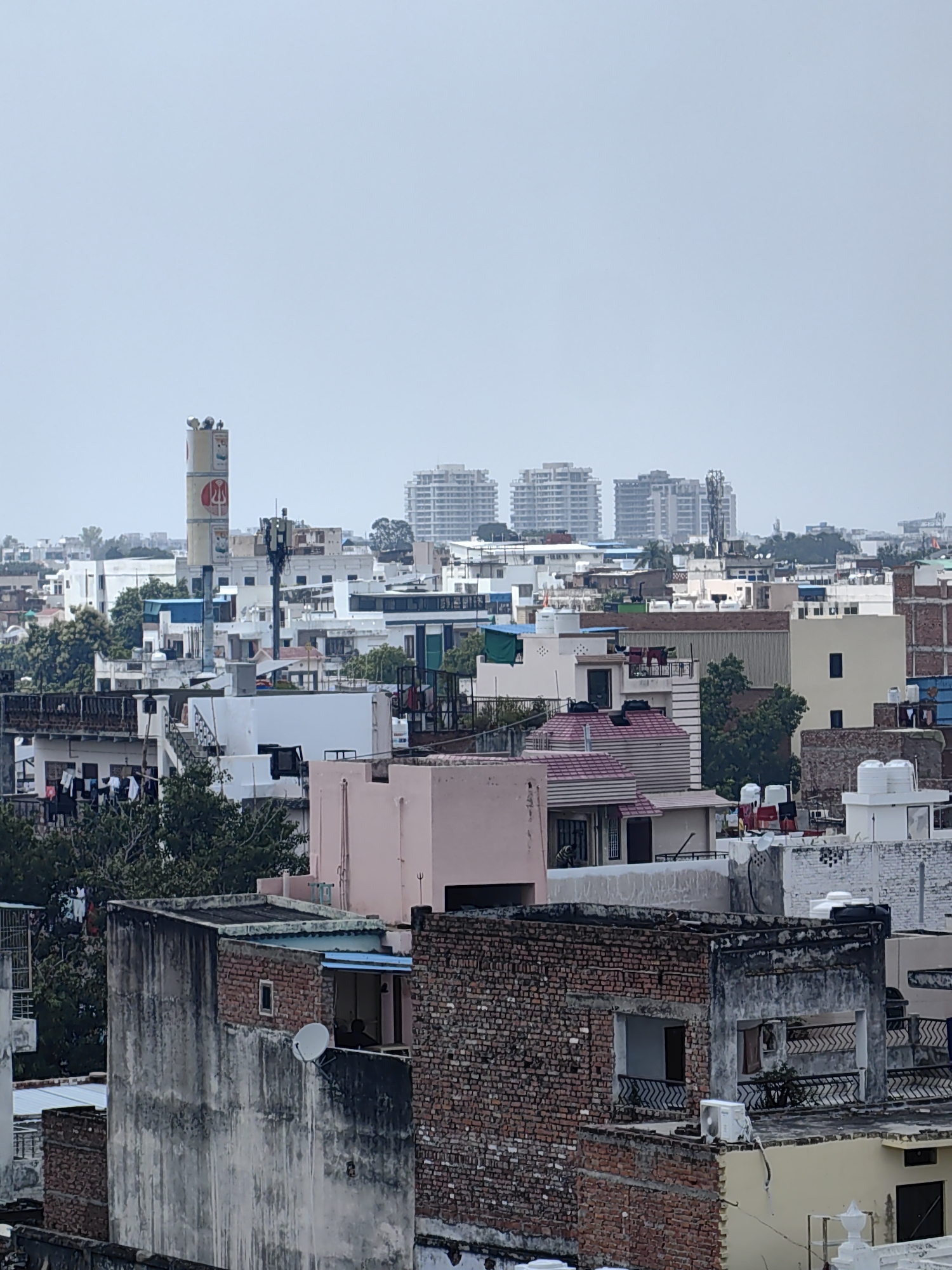 The image is an aerial view of a densely populated urban area in Varanasi, India, on a cloudy day. The foreground is dominated by the rooftops of various buildings, showing a mix of architectural styles and states of repair. Several structures are made of brick, some appearing weathered and aged, while others are newer constructions with smooth plaster finishes. Clothes are seen drying on lines on some of the rooftops.
Further into the scene, a tall, cylindrical structure, likely a water tank or a telecommunications tower, stands out among the buildings. It is adorned with circular logos, one of which contains an orange symbol. Multiple antennas and communication equipment are visible on top of a shorter tower next to it. Beyond this, a cluster of modern, multi-story buildings with repetitive window patterns rises against the hazy sky. The overall impression is of a busy, developing cityscape with both older and newer constructions coexisting.