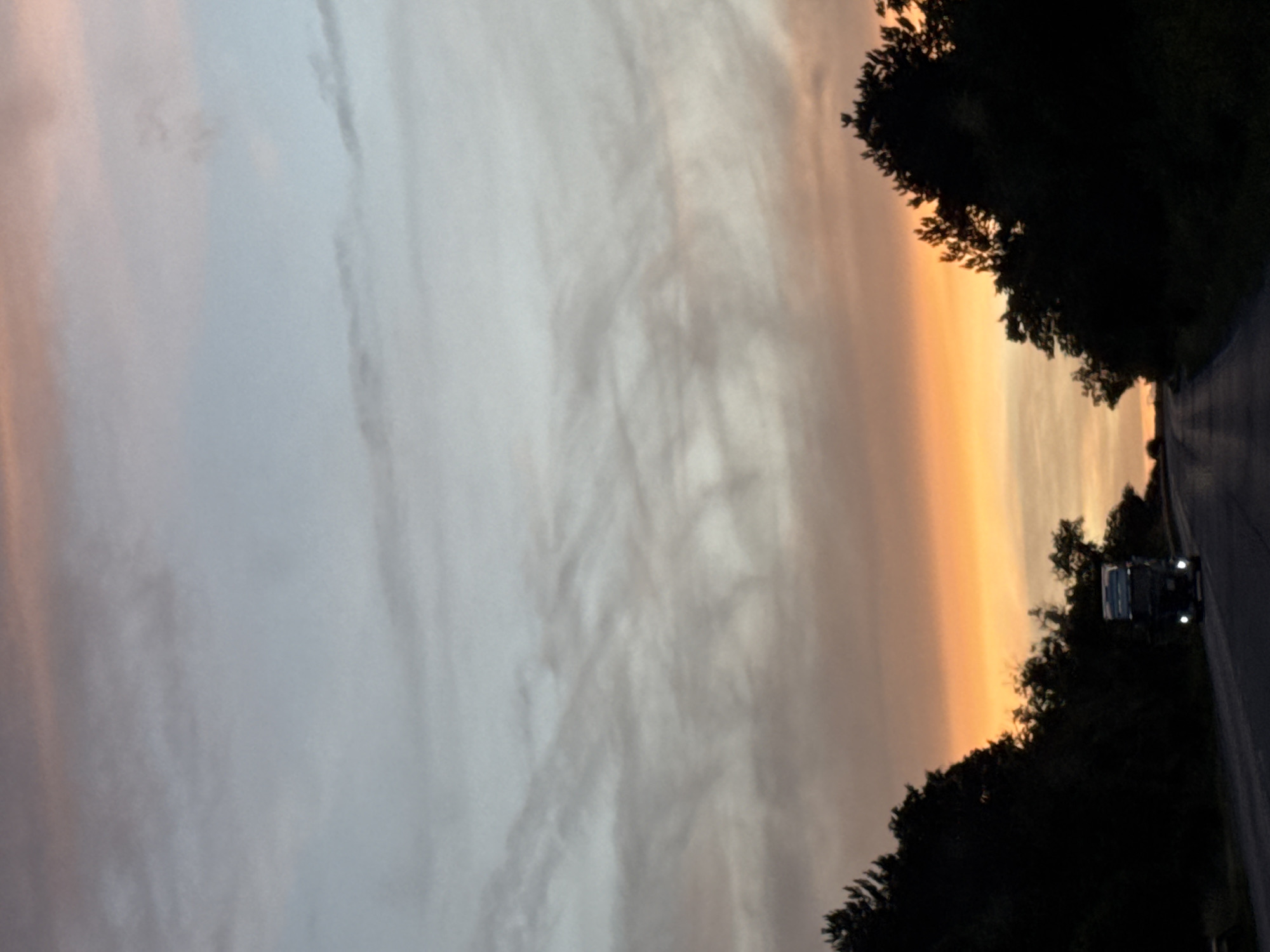 An asphalt road curves through a dark forested landscape under a twilight sky. A semi-trailer truck, illuminated by its headlights, drives away from the viewer on the road. The sky transitions from a pale blue near the top of the frame to a vibrant orange and pink glow near the horizon, with wispy clouds scattered throughout. The scene suggests travel during dusk or dawn, with the truck likely on a highway. The location is identified as Razvigorovo, Bulgaria.