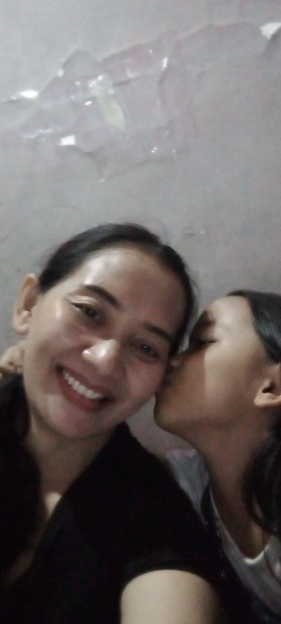 In a dimly lit indoor setting, a woman with dark hair is captured in a selfie-style photograph. She is smiling broadly, her teeth visible. To her right, a younger person with dark hair is leaning in and kissing the woman's cheek. The background appears to be a wall with a textured or damaged surface, possibly showing signs of water damage or peeling paint. The overall lighting is low, with some highlights on the woman's face. This image was taken in the city of Sumberwono, Indonesia.