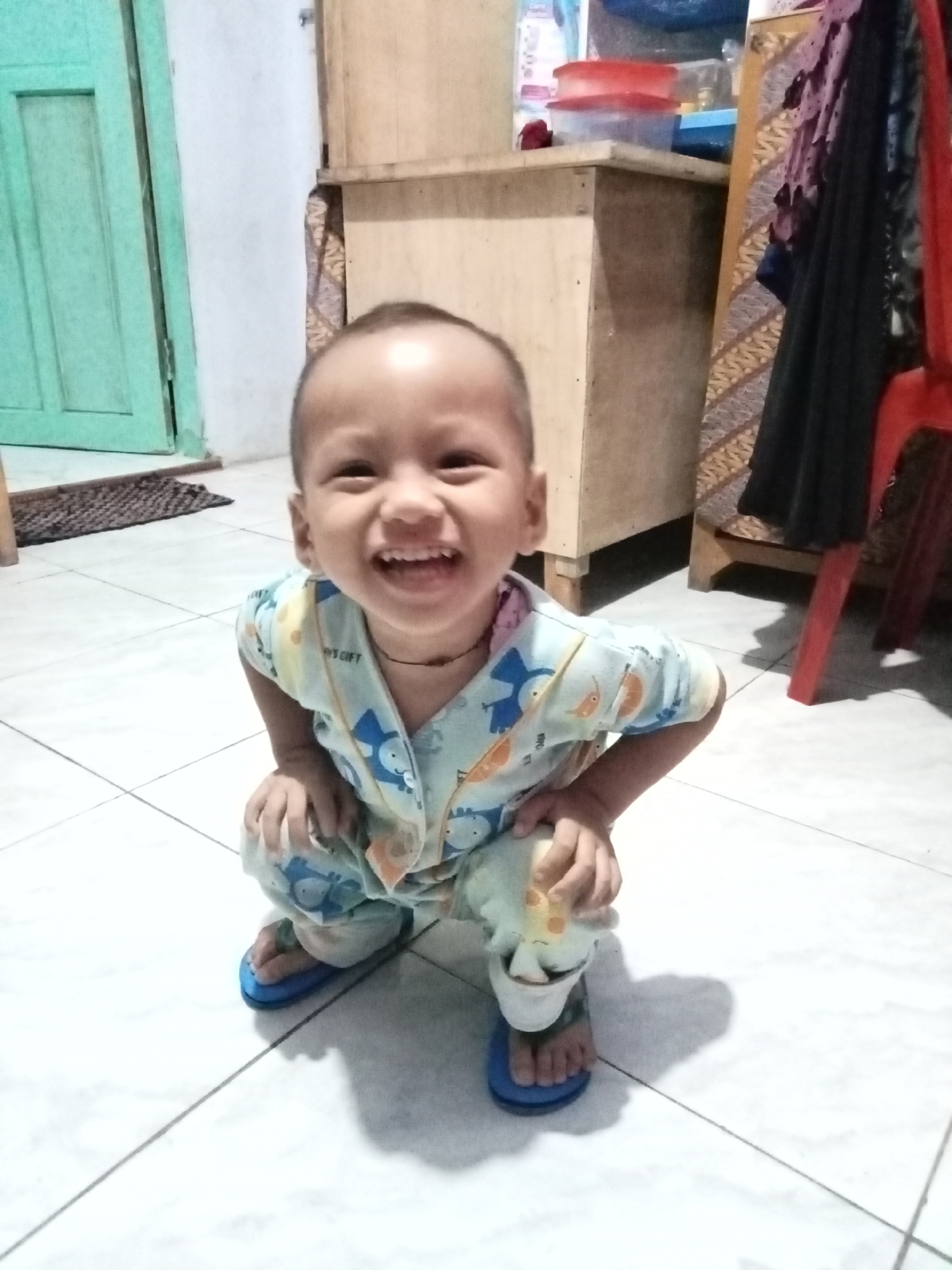 A young child, appearing to be between 2 and 4 years old, is photographed in a crouching position on a tiled floor. The child is wearing light blue pajamas with an all-over print of what appear to be cartoon animals and fruit. The child has a bright, wide smile, revealing teeth, and is looking directly at the camera. The child's head is shaved. The background includes a light-colored wall, a wooden cabinet with patterned fabric draped over it, and a green door. A red chair is visible to the right. The scene suggests an indoor setting, likely a residential area in Panyaungan Jaya, Indonesia, given the context. The lighting is somewhat dim, casting shadows on the floor.