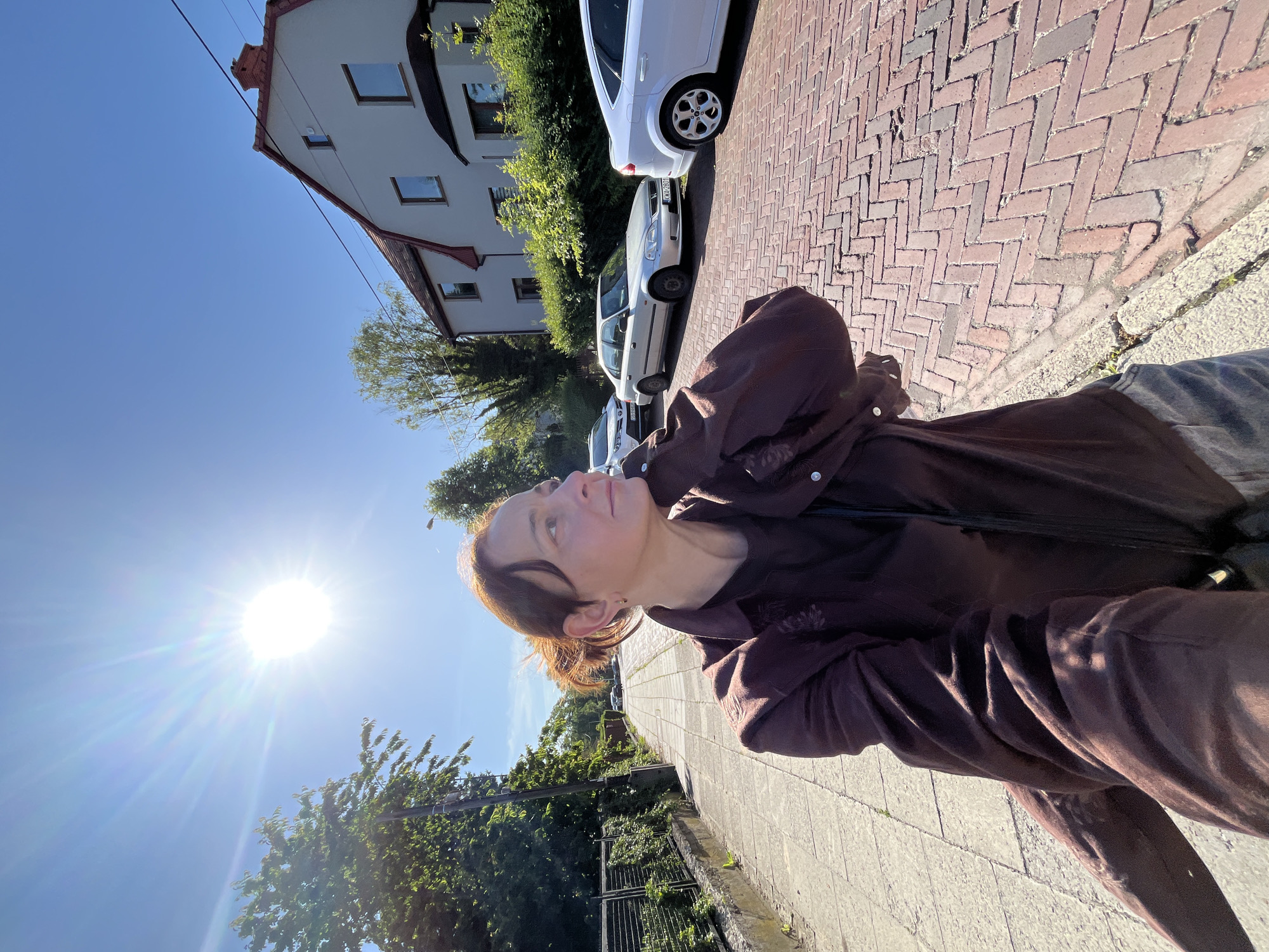A woman is taking a selfie on a street paved with red bricks. She is wearing a dark brown button-down shirt and gray pants. The sun is shining brightly in the clear blue sky, creating a lens flare in the image. To the left of the woman is a sidewalk made of concrete slabs, bordered by a metal fence and lush green foliage. To the right of the woman, several cars are parked along the brick street. In the background, a white house with a red tile roof is visible. The scene suggests a sunny day in a residential area.