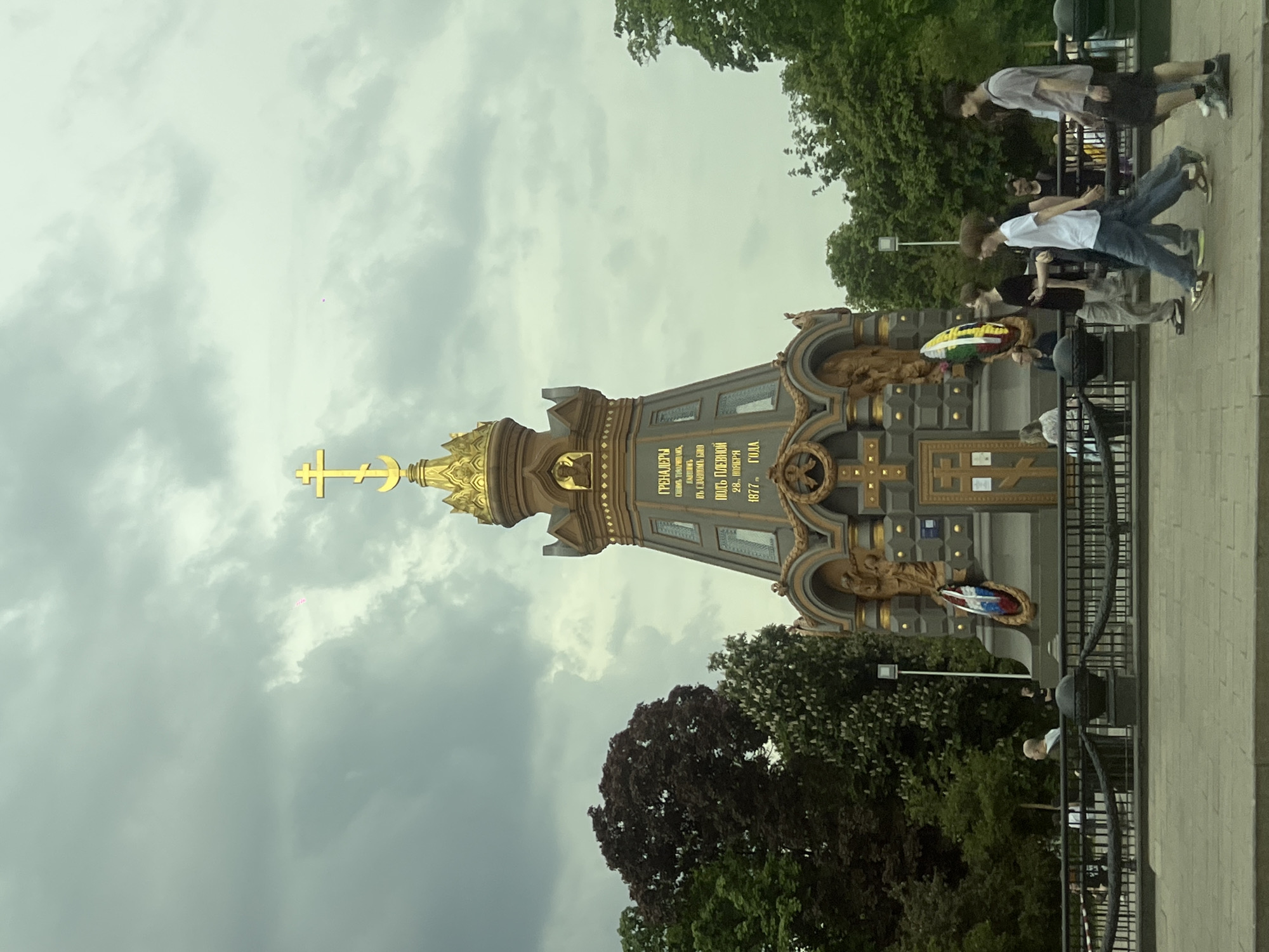 The image captures the Monument to the Grenadiers, located in Moscow, Russia. The monument features a tall, ornate structure with a golden cross on top, and text in Russian is inscribed on its surfaces. A group of people, some of whom are walking, can be seen in the background. The environment includes trees and a cloudy sky. A wreath with the colors of the Lithuanian flag hangs on the monument.