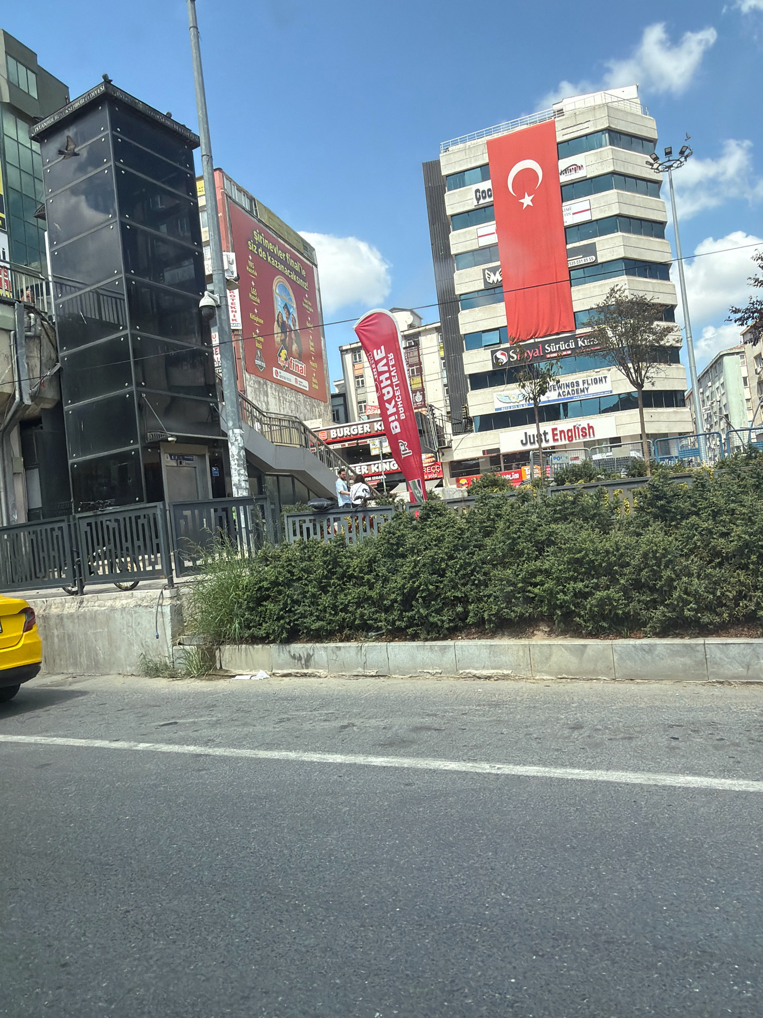 The image depicts a street scene in Bahçelievler, Turkey, dominated by a multi-story building flying a large Turkish flag. The building also features signage for "Suryal Sürücü Kursu" (driving school) and "Just English" language academy. To the left, a modern glass elevator structure is visible, with a pigeon in flight near the top. Adjacent to the elevator, a billboard with Turkish text and images is displayed. A red banner with white lettering, possibly an advertisement, is also present. In the foreground, a yellow taxi is partially visible on the left. The street is bordered by a manicured hedge and a sidewalk. The sky is blue with scattered clouds, and the scene appears to be illuminated by daylight. Several people are seen on a walkway or staircase in the middle ground.