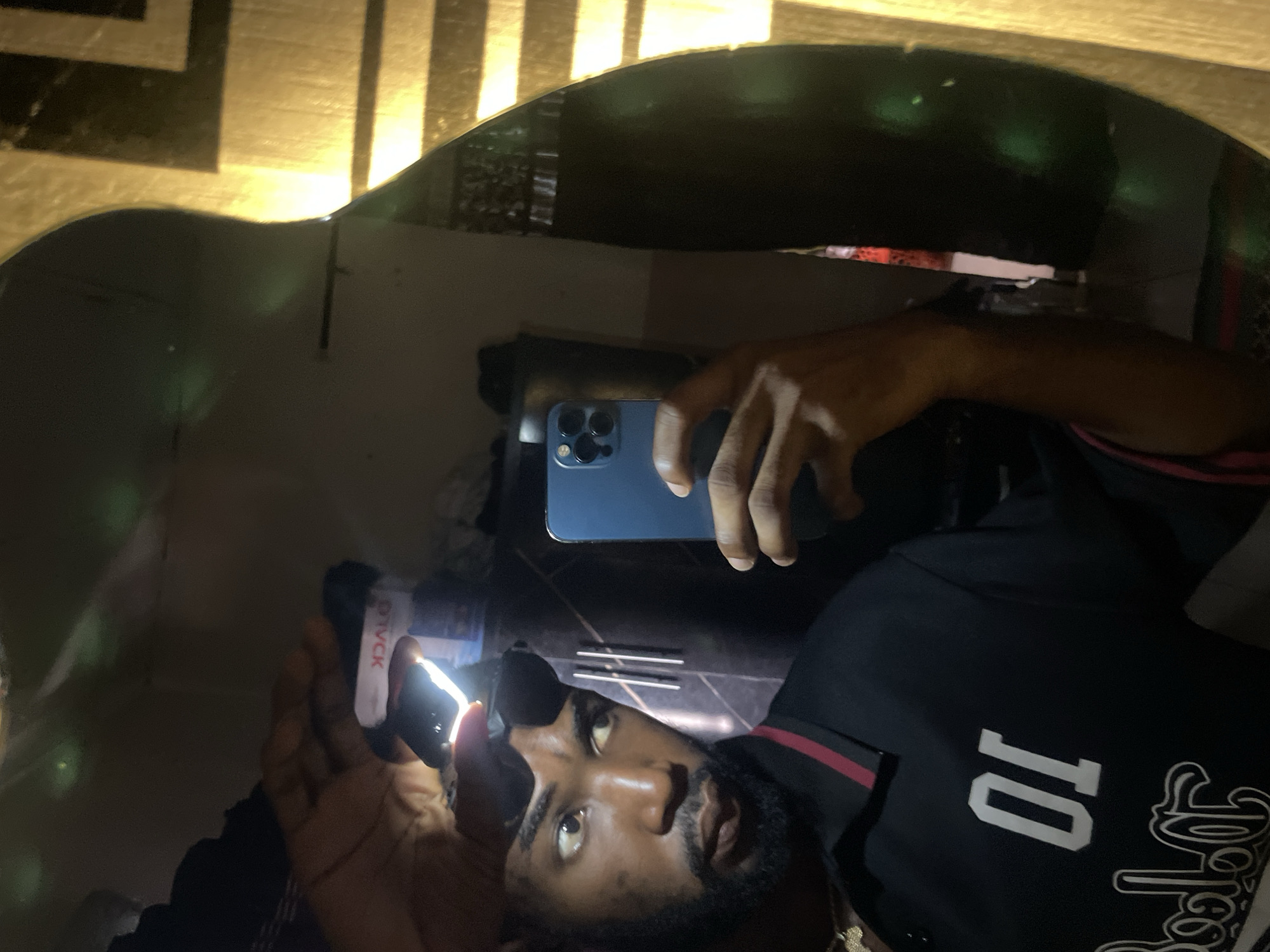 An adult male is depicted in a mirror selfie taken in an indoor setting within Lokoja, Nigeria. The mirror is irregularly shaped, resembling the outline of a guitar's body, and has a dark, reflective surface showing subtle green light reflections and minor surface imperfections. The man has a dark complexion, a well-maintained beard, and short dark hair. He is wearing a black polo shirt with a red and black striped collar, featuring the white number "01" and stylized white script below it on the front. A gold-colored chain with a pendant is visible around his neck. On his forehead, he wears a black headlamp actively emitting a bright white light, positioned above black-framed sunglasses that are pushed up onto his head. He holds a blue smartphone, identifiable as an iPhone due to its triple camera array, in his right hand, pointed towards the mirror. His gaze is directed slightly to the viewer's left. The background reflected in the mirror is dark, indicating low ambient light. Visible elements in the reflection include a white object with inverted red text "DYCK" in the upper portion, and a golden, horizontally striped surface (possibly blinds or a patterned wall) along the right edge of the mirror. Vertical metallic bars are also faintly visible behind the smartphone's reflection.
