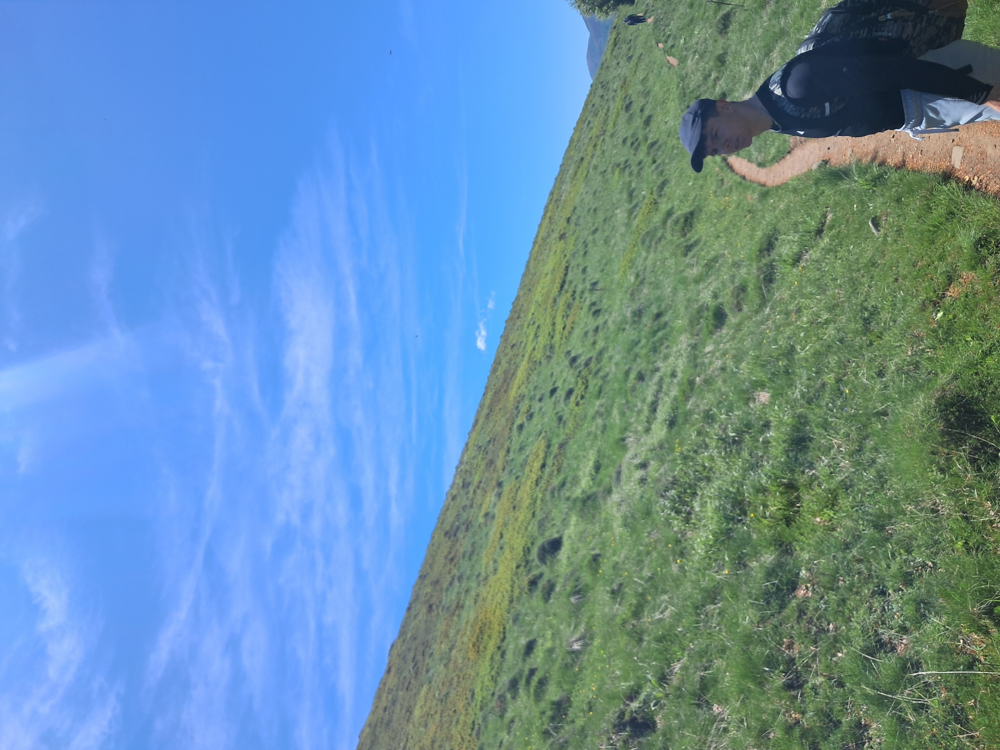 The image shows a grassy hillside under a bright blue sky with wispy clouds. A person is standing on a narrow dirt path along the upper edge of the hill. They are wearing a dark-colored top, light-colored shorts, a dark baseball cap, and a backpack. The grass on the hillside is mostly green with patches of yellow flowers. The photo was taken in Stokite, Bulgaria.