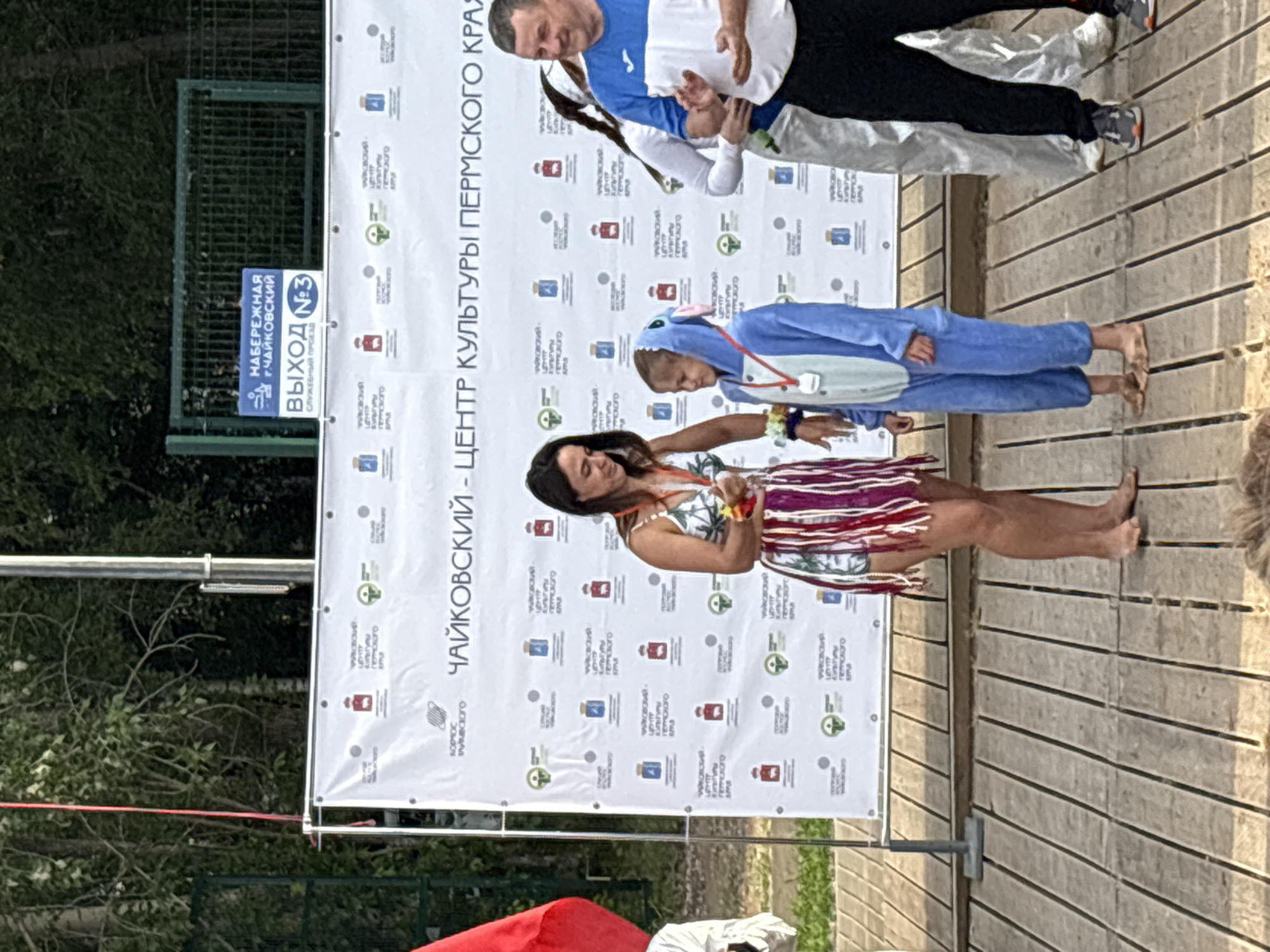 A woman in a colorful fringed dress stands on a wooden boardwalk next to a child in a blue Stitch costume. The woman is holding a medal or award in her left hand, presenting it to the child. Behind them is a large banner with Russian text, including "Чайковский - Центр культуры Пермского края" (Chaikovsky - Center of Culture of the Perm Territory). A man in a blue and white shirt stands to the right, clapping. In the background, a green fence and trees are visible. A blue sign attached to the fence reads "НАБЕРЕЖНАЯ г. ЧАЙКОВСКИЙ ВЫХОД №3" (Embankment of Chaikovsky, Exit No. 3). This scene appears to be an outdoor event, possibly a ceremony or festival, taking place at the Chaikovsky embankment.
