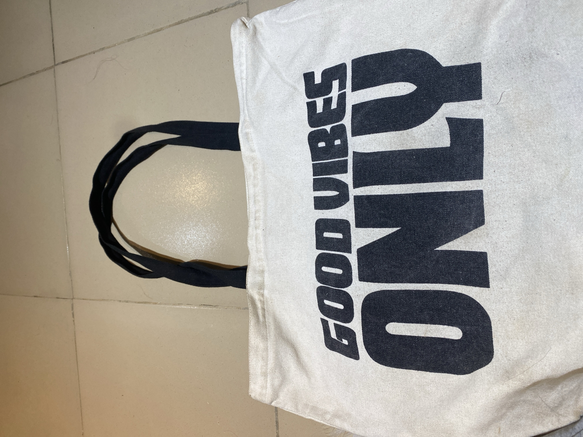 A close-up, vertically-oriented photograph shows a white tote bag with black handles resting on a beige tiled floor. The bag has the text "GOOD VIBES ONLY" printed on it in bold, blocky, black letters. The photo does not provide any information about the location being Jos, Nigeria or any people, actions, or interactions.