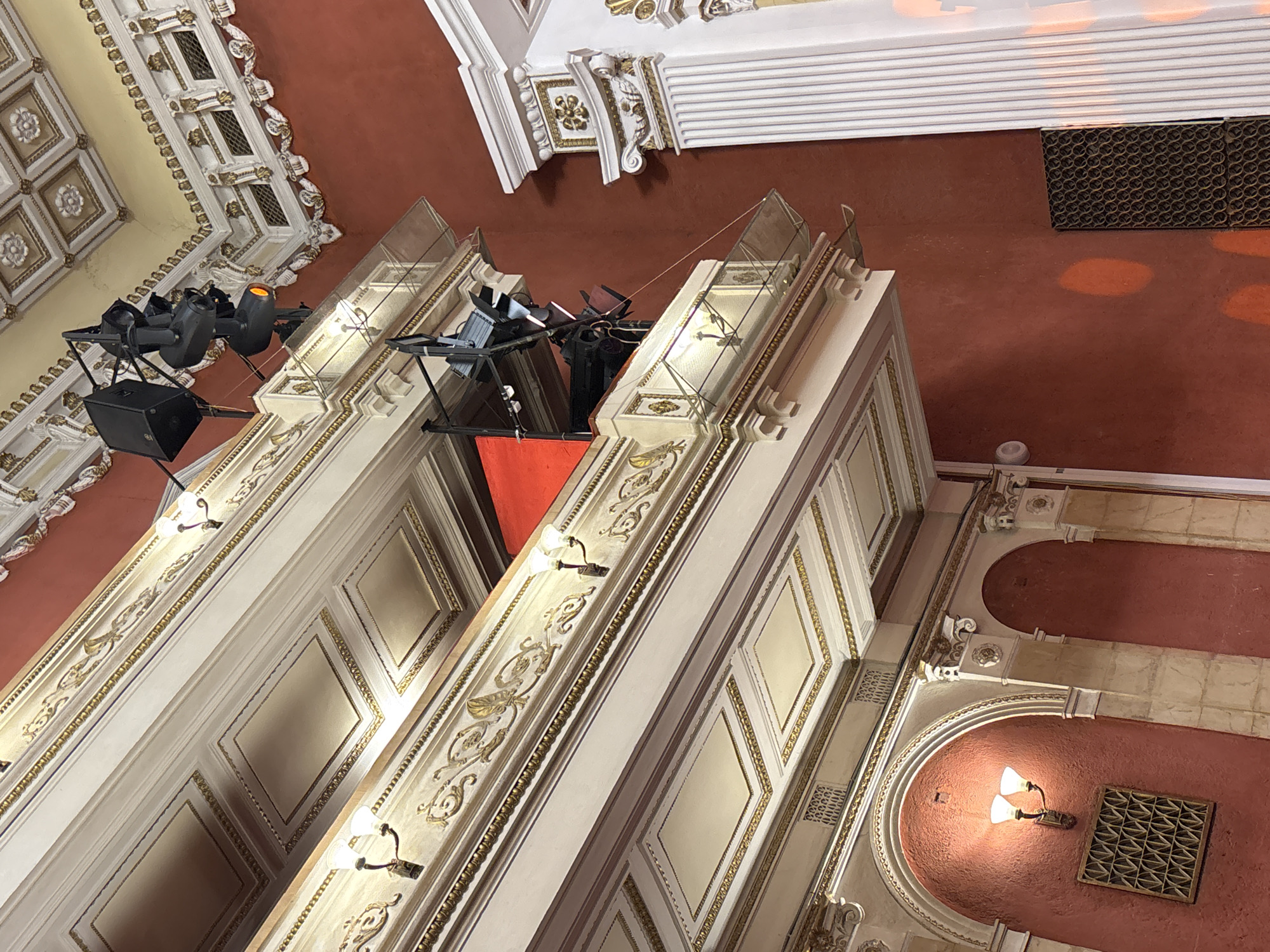 The image shows the interior of a theater in Perm, Russia. The ceiling is adorned with ornate designs in white, gold, and beige. Two prominent white pillars with gold trim and glass enclosures are visible, supporting lighting equipment. The floor and walls are a reddish-brown color with arched architectural details. A vent is noticeable in the floor/wall.