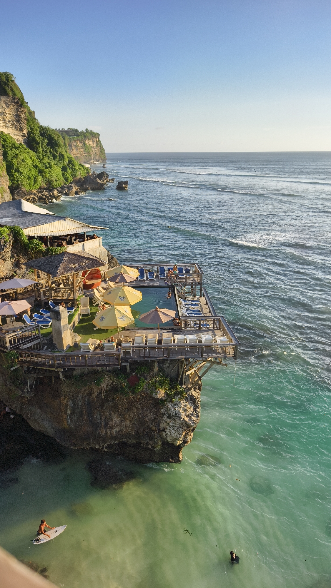 The image depicts a coastal scene in Pecatu, Indonesia, featuring a wooden structure built atop a rocky cliff. This structure houses a restaurant and a swimming pool area with lounge chairs and umbrellas. Several people are present: some relaxing on the lounge chairs, others swimming in the pool, and a surfer paddling in the turquoise water below the cliff. The ocean stretches into the horizon, with gentle waves. Lush vegetation covers the cliffside to the left of the structure. The sky is clear blue, suggesting a sunny day.