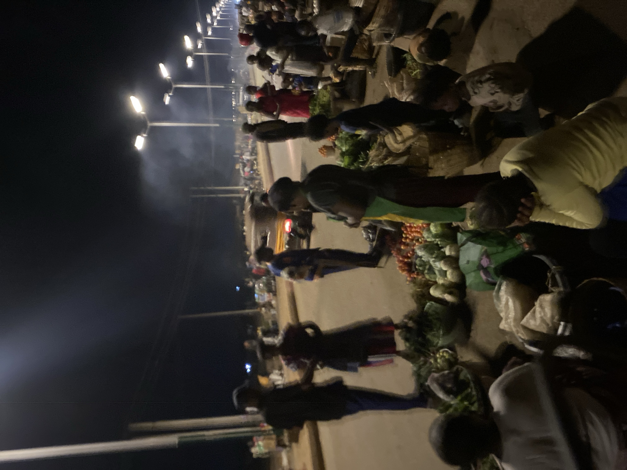 This vertical image depicts a busy outdoor night market in Bukuru, Nigeria, illuminated by a series of bright streetlights against a dark sky. Numerous people, predominantly adults, are visible throughout the scene, engaging in commercial activities. The market appears to be focused on fresh produce, with several stalls displaying piles of vegetables, including leafy greens, red spherical items (likely tomatoes), and other light-colored produce.
Many individuals are seen bending or squatting to interact with the goods on display, suggesting they are either vendors arranging their wares or customers examining products. The people are dressed in casual clothing. A distinct atmospheric haze or smoke is visible around the streetlights, adding to the ambiance of a bustling urban night market. The ground is a mix of paved and unpaved surfaces. The overall scene conveys a lively and active marketplace under artificial light after sunset. No discernible text is visible in the image.