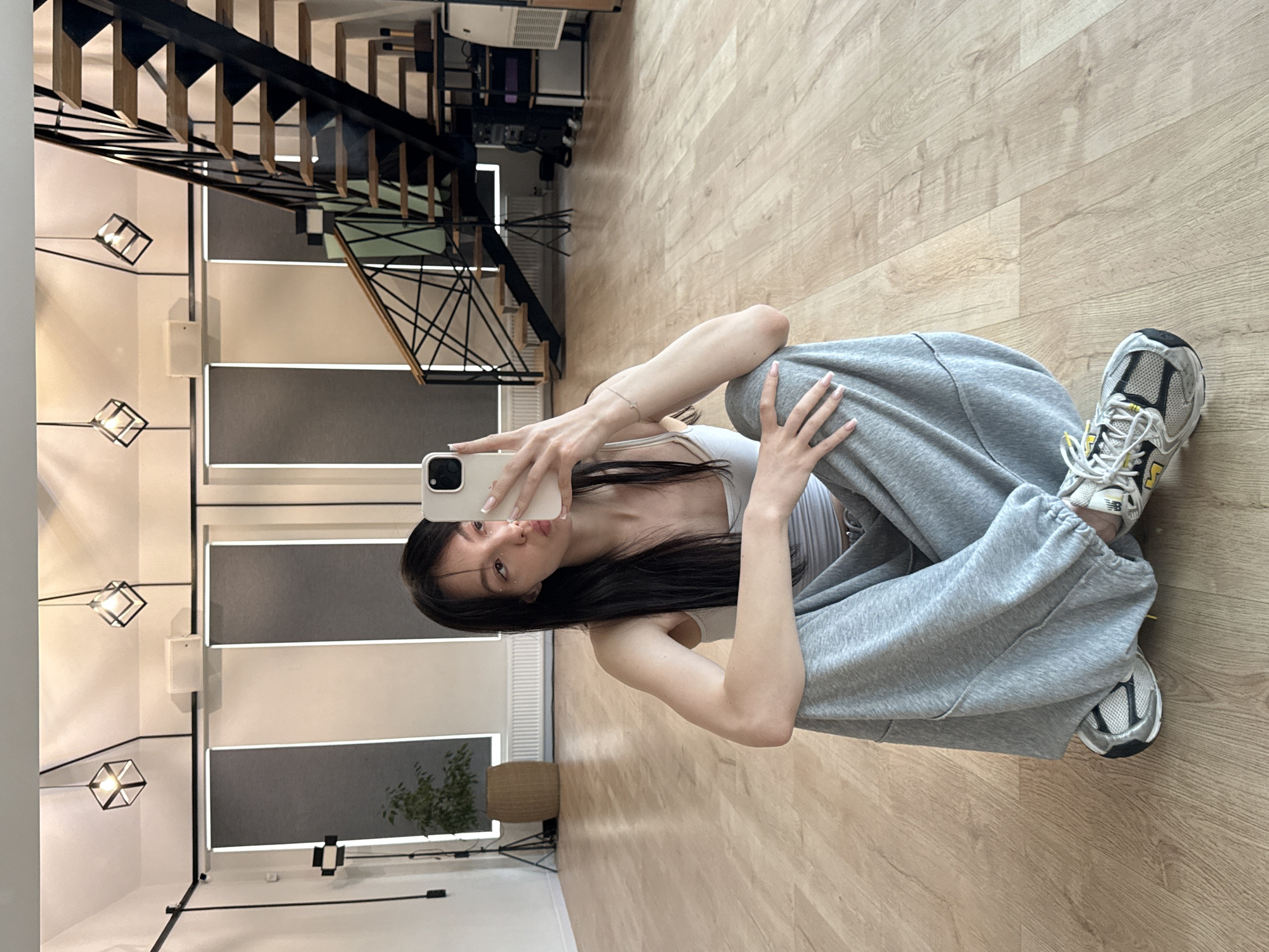 The media file is a vertical, full-body, mirror selfie taken by a young woman indoors. She is sitting on a light-colored wooden floor, with her knees bent and her legs crossed. The woman has long, dark hair and is wearing a light gray tank top and loose-fitting gray sweatpants. She is holding a white smartphone in her right hand, angled to capture her reflection. Her left arm rests on her leg. She is wearing white and gray athletic shoes with yellow accents.
The setting appears to be a modern interior with a staircase in the background. The staircase has black metal railings and wooden steps. There are geometric light fixtures hanging from the ceiling. The walls are a mix of white and gray panels, with some containing integrated lighting. A plant in a basket and a tripod are visible on the floor in the mid-ground. The overall ambiance is casual and relaxed.
The image is well-lit, suggesting it was taken during the daytime or under bright indoor lighting. There is no visible text in the image. The woman's expression is playful, with a slight pout. The photo's composition emphasizes the woman and her outfit, with the background providing context for the indoor environment. The location context of Zakharovo, Russia, does not provide any immediately discernible visual cues within the image itself.