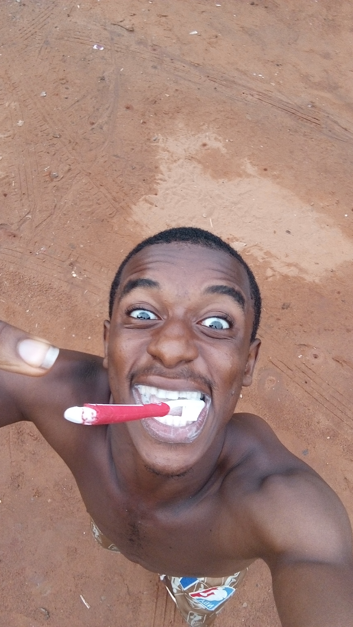 A dark-skinned man with short black hair is smiling widely while brushing his teeth with a red and white toothbrush. His eyes are wide open and light blue. He is shirtless and wearing patterned shorts. The background is reddish-brown, sandy soil. There is no visible evidence to determine the location is in Onitsha, Nigeria.