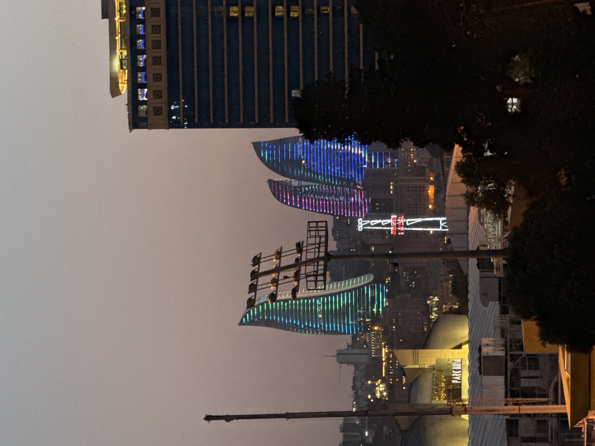 The image depicts the Flame Towers in Baku, Azerbaijan, at dusk. The Flame Towers, three skyscrapers shaped like flames, are illuminated with vibrant blue and green lights, creating a striking visual against the twilight sky. To the right of the Flame Towers, a tall, dark blue building with horizontal windows is visible. In the foreground, a large dark tree partially obstructs the view. Closer to the bottom of the frame, a building with the text "PARK BULVAR" is illuminated in yellow. There are no visible people in the image. The overall scene is a cityscape at the transition from day to night, with artificial lights becoming prominent.