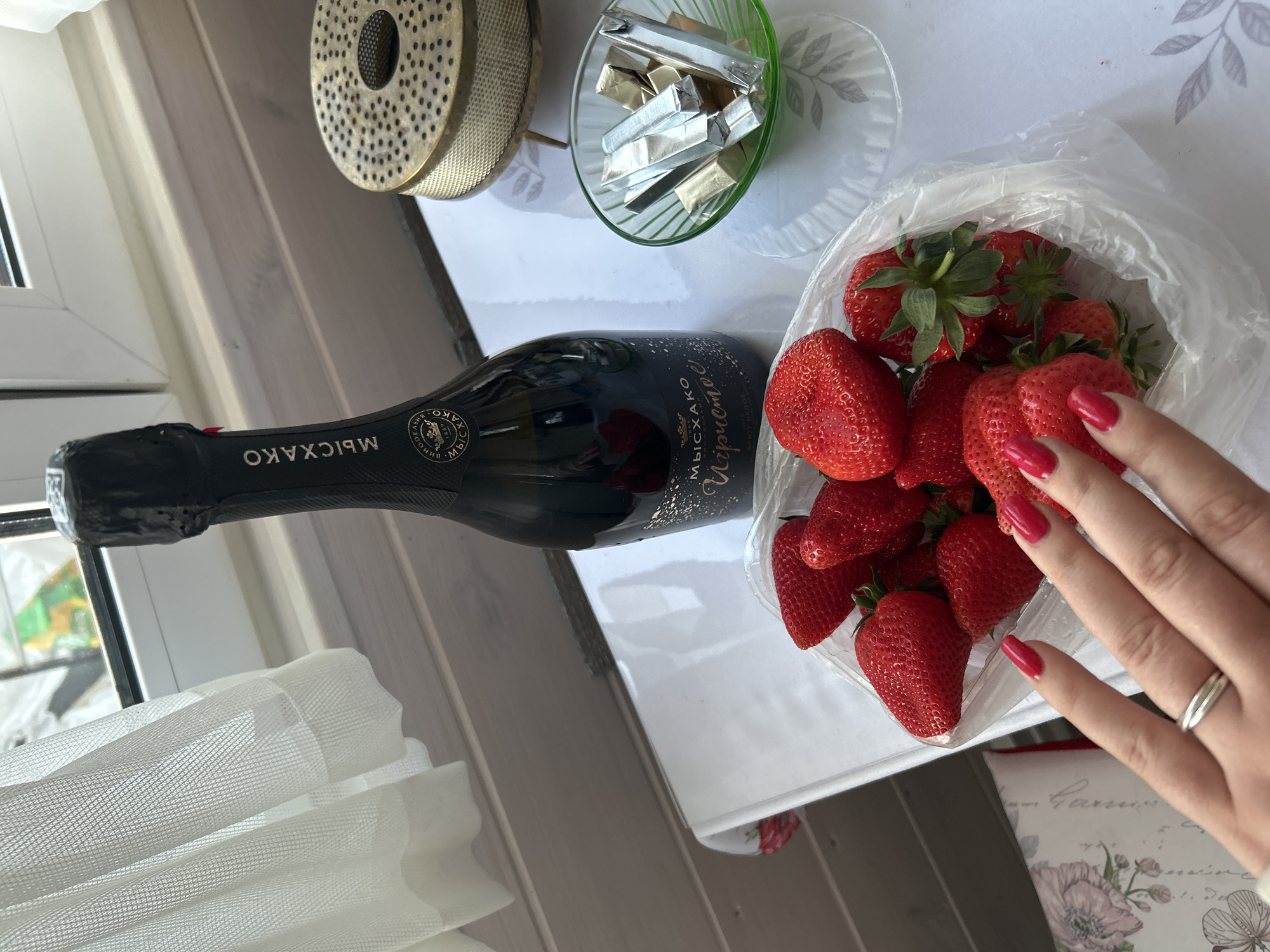 On a table in Bashur, Russia, a still life is arranged. A bottle of sparkling wine labeled "Myskhako Igristoe" lies next to a clear plastic container filled with strawberries. A woman's hand with red nail polish is reaching into the strawberries. A small green glass bowl containing foil-wrapped candies is positioned next to a cylindrical metal object with a perforated top. A white sheer curtain is visible in the background near a window. The table is white.