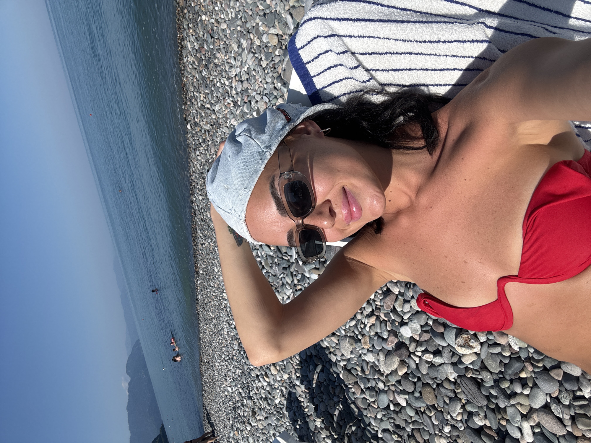 A woman, wearing a red bikini top and a denim cap, lies on a beach chair on a pebble beach. She is wearing sunglasses and has her left arm behind her head. The sea stretches out behind her, with mountains in the distance. The scene is set in Batumi, Georgia, evidenced by the distinctive mountainous coastline characteristic of the region. Several other people are visible in the water, some swimming and one on an inflatable ring.
