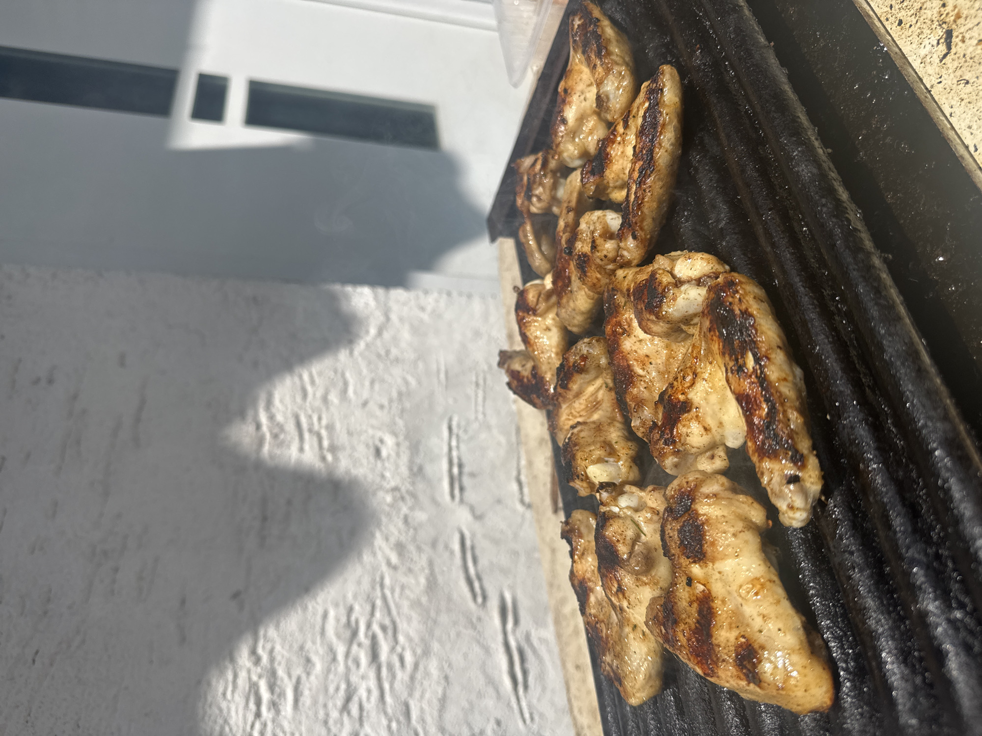 The image depicts approximately a dozen chicken wings grilling on a black, ridged grill pan outdoors under strong direct sunlight in Hattersheim am Main, Germany. The chicken wings exhibit golden-brown coloration with distinct char marks, indicating active cooking, and faint wisps of steam are visible rising from the pan. The grill pan rests on a light-colored, possibly stone or concrete, surface. In the background, a white, textured wall, likely stucco or rough plaster, is prominently visible, displaying sharp, elongated shadows from an unseen overhead structure or foliage, consistent with direct sunlight. To the right of the frame, a segment of a white door or window frame with a dark vertical element is discernible. A transparent plastic container is partially visible in the upper right quadrant of the image. No individuals are present in the scene.