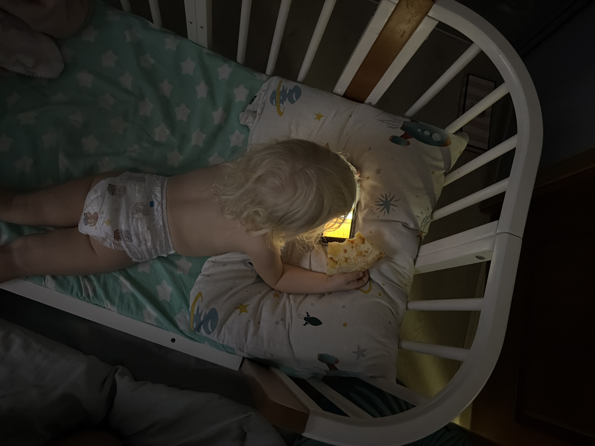 An overhead, low-light photograph captures a young child, likely a toddler, lying on their stomach inside a white wooden crib. The child has blonde, curly hair and is wearing a white disposable diaper decorated with cartoon characters. They are resting on a white pillow adorned with space-themed illustrations of rockets, stars, and planets, over a light green sheet patterned with white stars.
The primary light source in the dimly lit room is a brightly glowing electronic screen, possibly a smartphone or tablet, which the child holds with both hands. This screen casts a warm yellow light onto the child's head, hands, and the pillow. Simultaneously, the child is holding and appears to be eating a slice of pizza with one hand.
The activity takes place indoors at night, suggested by the overall darkness of the scene, with the electronic device being the only significant source of illumination. The crib features white wooden slats and a curved end. While the image contains no specific visual cues to its location, this scene is set in Sochi, Russia. There is no visible text in the image.