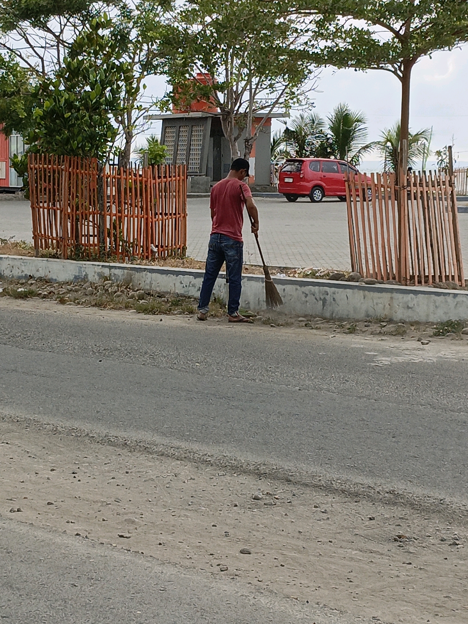 A man is sweeping the side of a road in Bulukumba, Indonesia. He is wearing a red t-shirt and blue jeans. He is holding a broom and sweeping debris from the curb. In the background, there is a red car, palm trees, and a fence. The road appears to be paved, and there is gravel on the side of the road. The sky is cloudy.