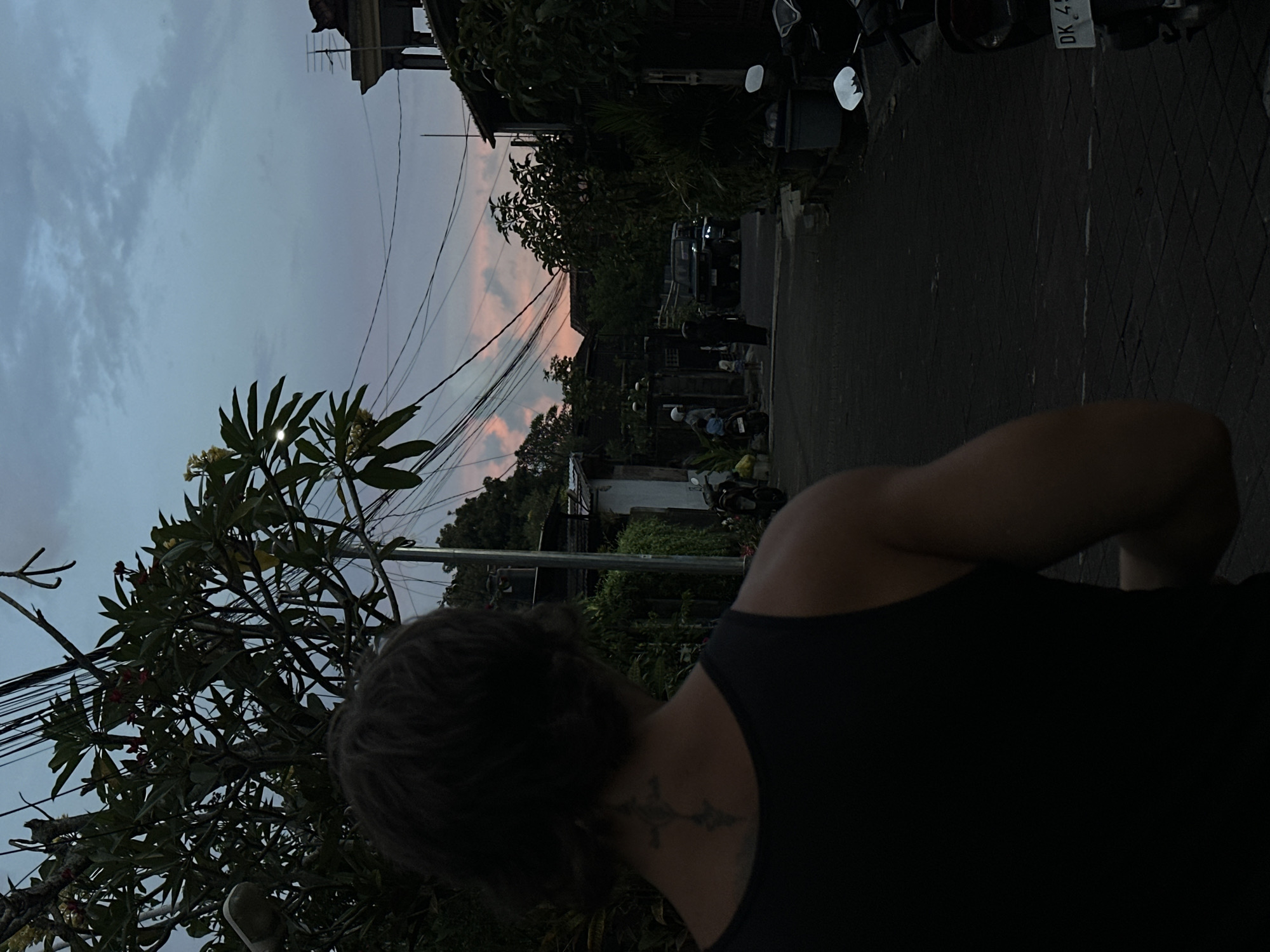 The image, taken in Dalung, Indonesia, shows a person wearing a black tank top from the back, with a tattoo visible on the neck. The scene is an urban street with a narrow paved road and buildings obscured by lush tropical foliage. Overhead power lines stretch across the scene against a dusky sky with muted pink highlights, suggesting either sunrise or sunset. Motorcycles are parked along the side of the road. Several people are further down the road. The lighting is dim.