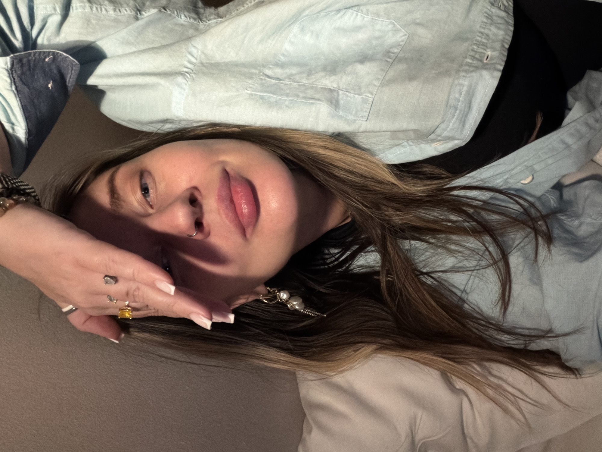 A young woman with brown hair is reclining, wearing a light blue button-up shirt. She has rings on her fingers and an earring in her left ear. Her left hand is resting on her forehead. No information in the image suggests the location is Moscow, Russia. The background appears to be a plain wall and a white pillow or bedding.