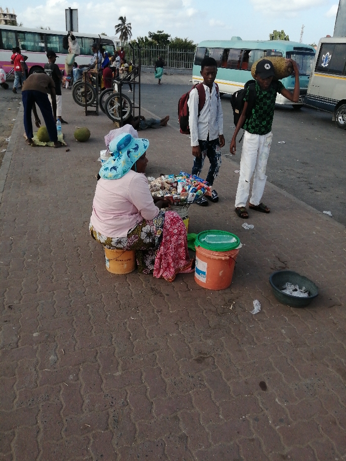 A woman sits on a yellow bucket on a brick sidewalk, selling snacks from a silver container. She wears a pink shirt, a floral skirt, and a blue hat. Beside her are two orange buckets and a bowl filled with white packets. Two young men stand nearby. One carries a backpack and wears a white shirt and patterned jeans. The other carries a heavy object on his shoulder. In the background are buses, people, and carts with wheels. Someone is lying on the ground near the carts.