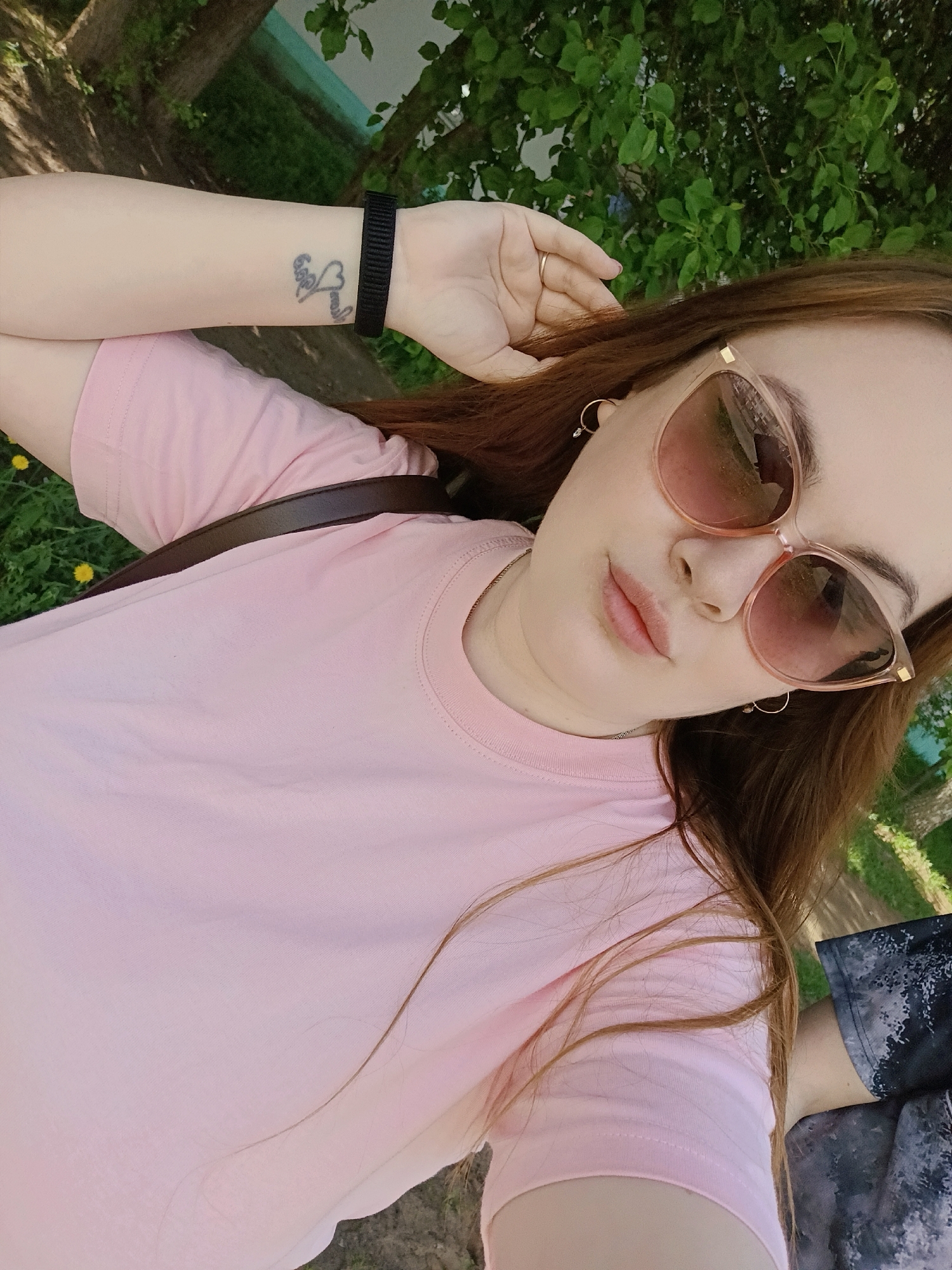 The photograph captures a young woman from a high-angle perspective in what appears to be an outdoor setting. She is wearing a light pink T-shirt and sunglasses with a light brown tint. Her hair is long and auburn in color. There is a dark-colored wristband and a tattoo visible on her left forearm. The environment includes foliage of green trees and grass and is located in the city of Chaikovsky, Russia.