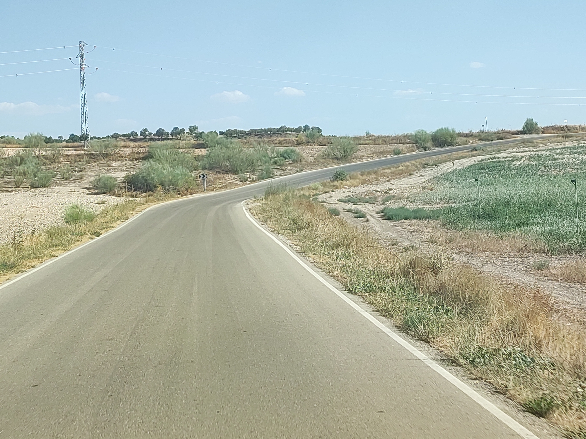 The image shows a paved road curving to the left in a rural landscape under a clear blue sky with a few wispy clouds. To the left of the road, there is dry, light-colored soil with sparse, dry vegetation and a few small green bushes. Further in the background to the left, a tall electrical pylon with multiple wires stretching across the scene is visible. Beyond the pylon, there are more trees and bushes, and the terrain appears to be hilly. To the right of the road, there is an area with dry grass and some cultivated green crops. The road itself is grey asphalt with a white line marking the right edge. A black and white curved arrow sign is visible on a post to the left of the road, indicating a sharp curve ahead. The scene appears to be a sunny day in a dry, semi-arid environment, likely in or near Illana, Spain, given the vegetation and terrain. There are no people or animals visible in the image.