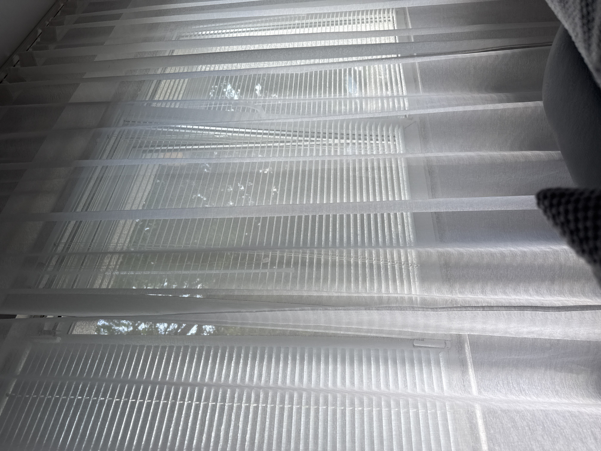 The image shows a view through a window covered with white, semi-transparent blinds. The blinds are partially open, allowing some light and a view of trees and foliage outside. A gray object, likely a piece of furniture, is partially visible on the right side of the frame. The location is not identifiable as Lovech, Bulgaria, based on visual information in the image.