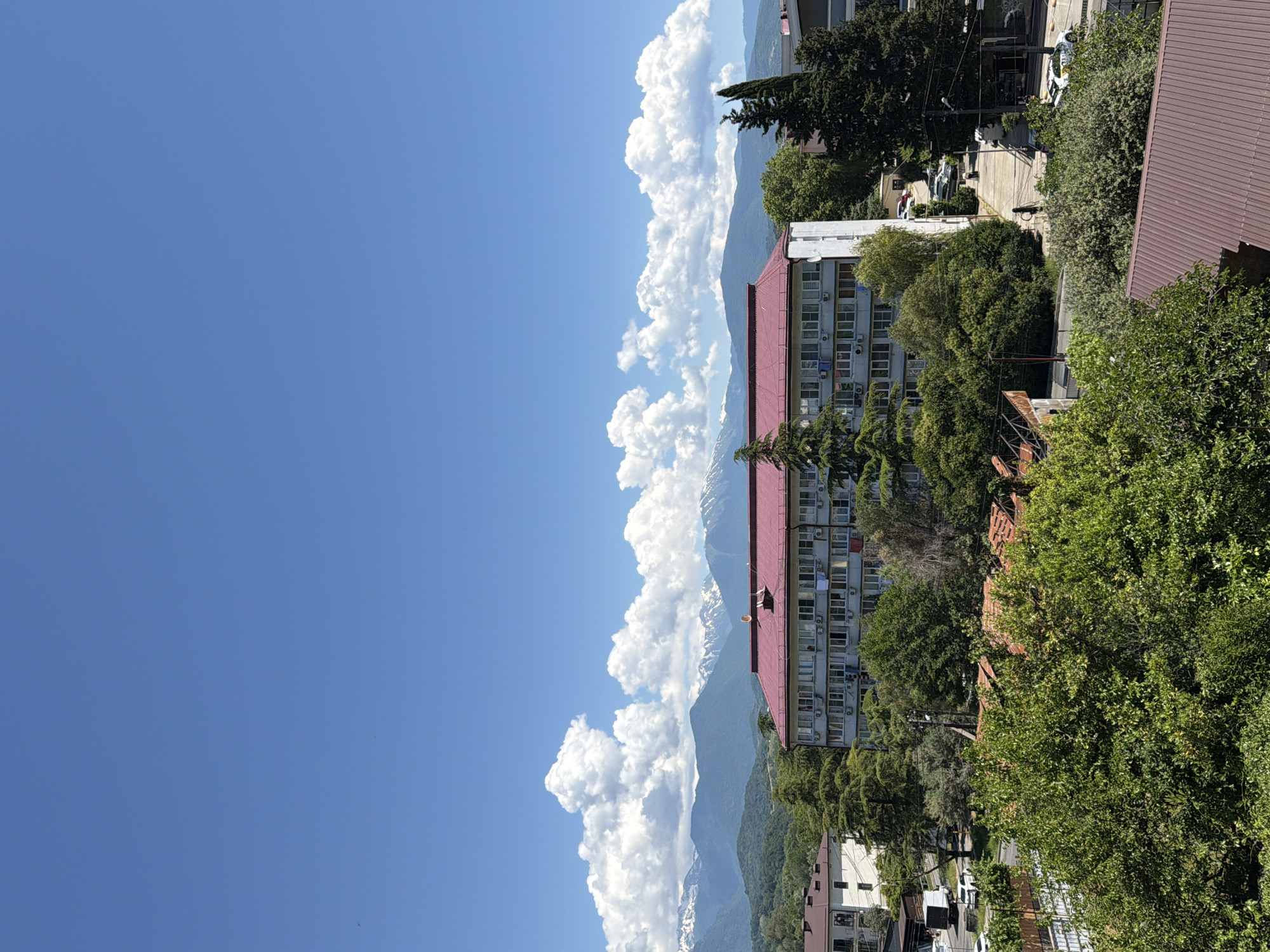 This is a vertical, high-angle, medium shot of a residential building in Sochi, Russia, on a sunny day. The building is light gray with a red roof and is surrounded by lush green trees. In the background, snow-capped mountains are visible under a bright blue sky with fluffy white clouds. The street in front of the building is lined with parked cars and a white truck. The scene is peaceful and serene, with no people clearly visible.