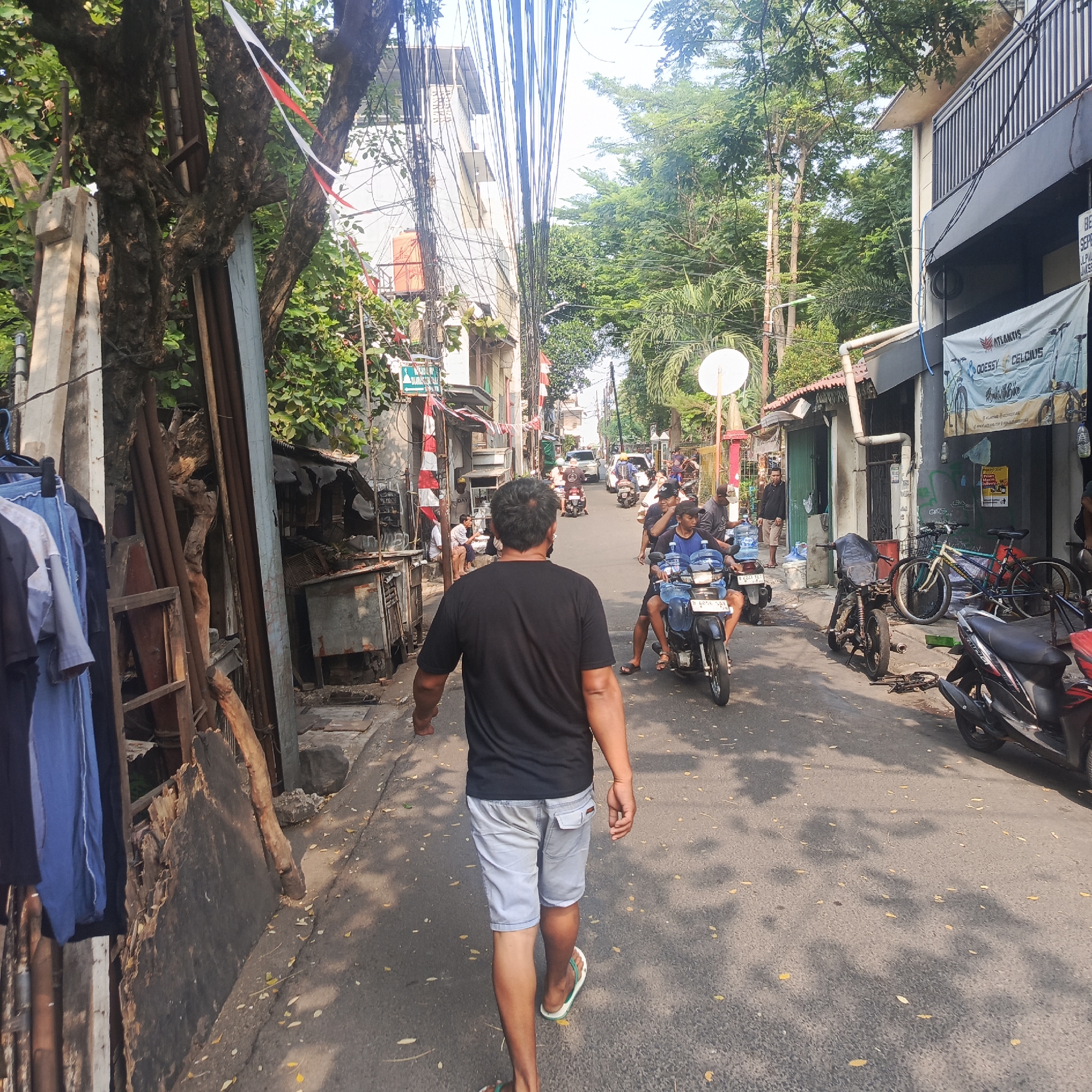 This is a medium-wide shot of a street scene in South Jakarta, Indonesia, likely in a residential or mixed-use area with narrow roads. The viewpoint is from behind a man walking away from the camera, towards the center of the street where traffic is moving. The man is wearing a black t-shirt and light grey shorts, and appears to be in his late 30s or 40s. He is wearing flip-flops.
The street is paved and appears somewhat worn, with scattered leaves and shadows cast by trees and overhead wires. On the left side of the street, there is a tree with a rough, gnarled trunk adorned with red and white Indonesian flags, indicating a national holiday or celebration. Adjacent to the tree are various items, including what appears to be scrap wood, metal poles, and hanging clothes.
To the right of the street, there are buildings of varying architectural styles. Some have balconies with railings, while others appear to be smaller shops or dwellings with corrugated roofs. Several motorcycles are visible on the street. One motorcycle in the foreground is ridden by two young men, one of whom is holding a large blue water jug. Another motorcycle, a scooter, is parked on the right side of the street. Further down the street, more motorcycles and a car are present, indicating moderate traffic. The overall atmosphere is that of a bustling urban environment, with a mix of residential and commercial elements. The presence of the flags suggests a local cultural event or observation.