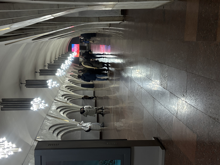 This image depicts the interior of the Yeritasardakan Metro Station platform in Yerevan, Armenia. The scene features a long, vaulted passage with a high ceiling and repeating series of decorative, light-colored architectural arches along both walls. A linear arrangement of dark, cylindrical chandeliers with multiple bright lights hangs from the ceiling, illuminating the space and casting reflections onto the polished reddish-brown granite or terrazzo tile floor.
Approximately 10-15 individuals are present on the platform. In the left middle ground, one person in a long white hooded coat and backpack holds a phone near their head, potentially taking a photo or making a call. Adjacent to them, two other individuals stand, appearing to converse. Further into the middle ground, a cluster of about five to six people in various winter coats and jackets stands together, seemingly engaged in conversation or waiting. Additional individuals are visible further down the platform towards the distant end.
In the left foreground, a vertical, dark gray digital information kiosk or advertising display stands, partially obscuring the left wall. A QR code is visible on its lower screen portion. At the far end of the platform, a large, brightly illuminated digital display screen is visible, exhibiting text that includes "BRAday" with a red background and a white heart graphic. The platform appears clean and well-maintained.