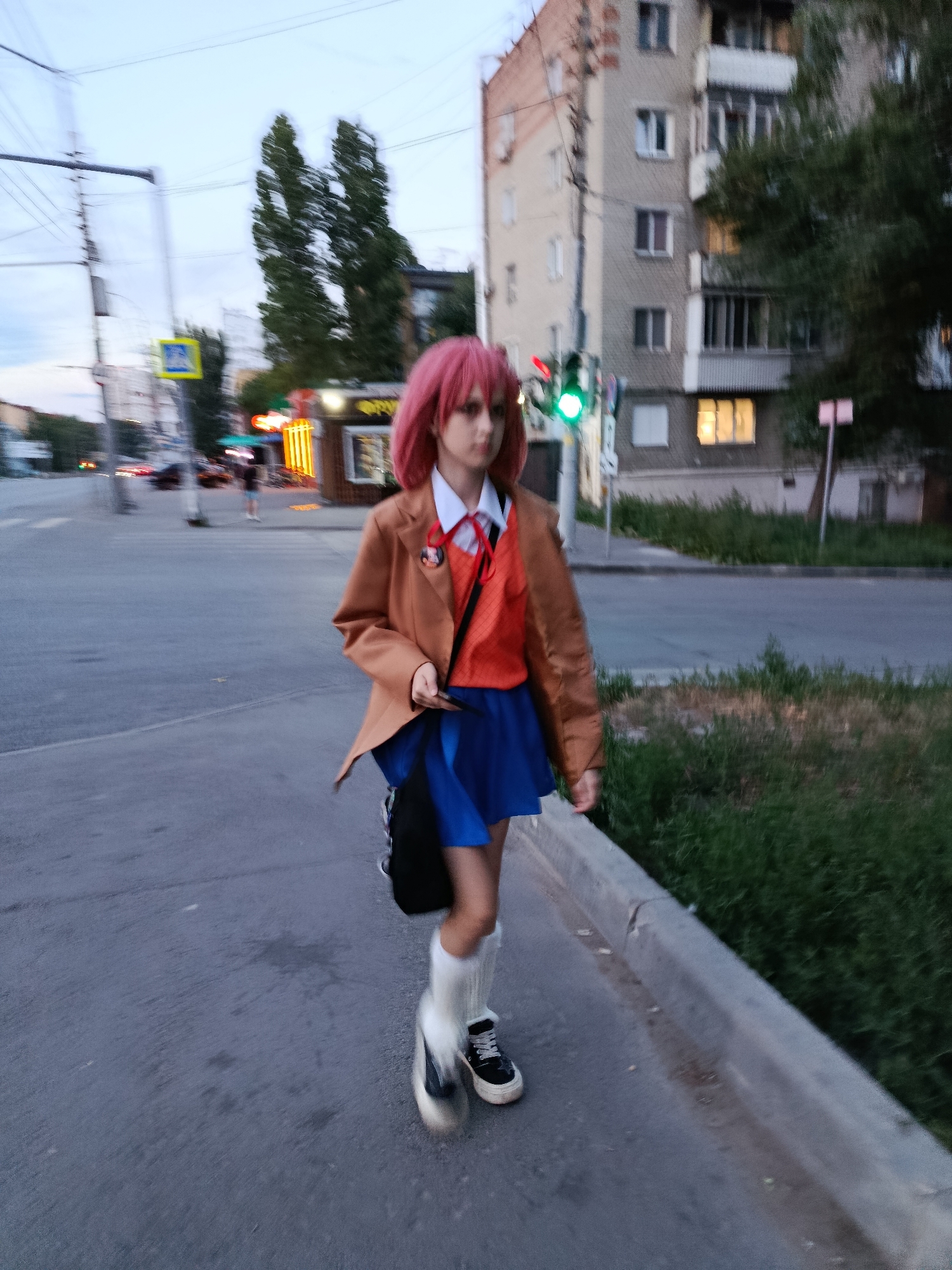 A person with pink hair, dressed in a brown blazer, orange shirt, blue skirt, white socks, and black sneakers, walks on a paved sidewalk. The person carries a black bag over their shoulder. They appear to be at a street intersection in Saratov, Russia. Traffic lights are visible, with a green light illuminated. Buildings, trees, and power lines are in the background. The lighting suggests it is dusk.