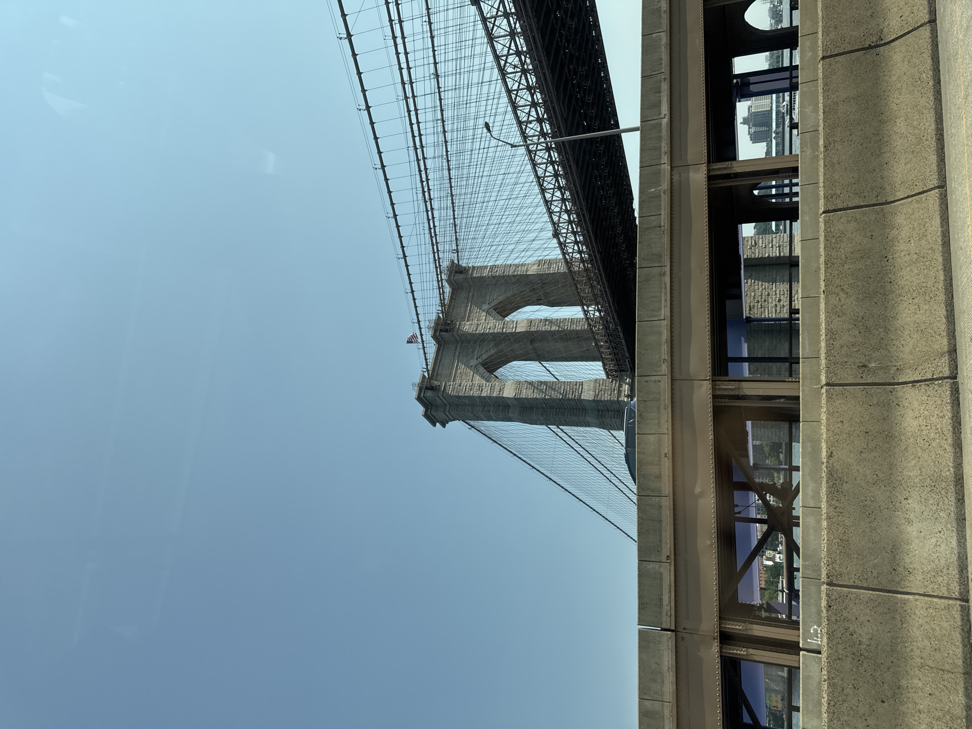 The image captures a daytime view of the Brooklyn Bridge in New York City, United States of America, seen from an elevated perspective, likely from a vehicle on an adjacent road. The sky is clear and blue, dominating the upper portion of the frame. The iconic stone towers of the Brooklyn Bridge are visible, with their Gothic arches and intricate suspension cables stretching across the sky. The bridge's roadway, a dark metal structure, is suspended below the cables. In the foreground, a concrete barrier and a section of a metal and glass structure, possibly part of an overpass or elevated roadway, obstruct the lower part of the view of the bridge. Through the windows of the foreground structure, glimpses of the water and buildings on the opposite shore can be seen. An American flag is flying from one of the bridge towers. No people are clearly discernible in the image. The overall scene conveys a sense of urban infrastructure and monumental architecture.