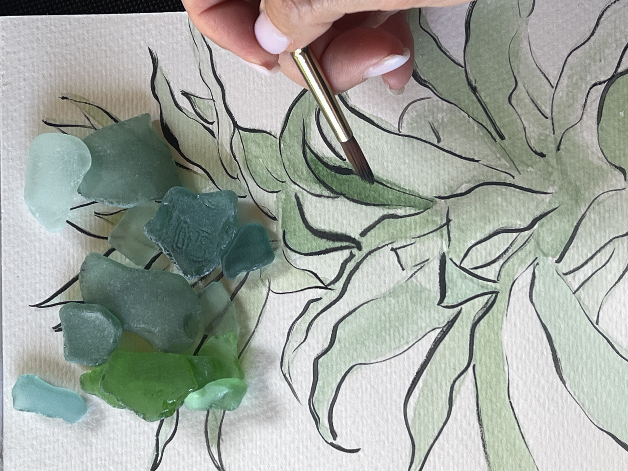 A close-up, overhead shot shows a hand holding a paintbrush over a watercolor painting of green leaves with black outlines. Several pieces of sea glass in varying shades of green and blue are scattered on the paper above the painting. The sea glass pieces are irregularly shaped and translucent. The watercolor painting depicts a stylized plant with broad, curving leaves. The hand is holding a thin paintbrush with a dark brown tip, applying a light green wash to one of the leaves. The background is a textured, light-colored surface. The overall scene suggests an artistic process, possibly creating a mixed-media artwork. The location is not specified in the image.