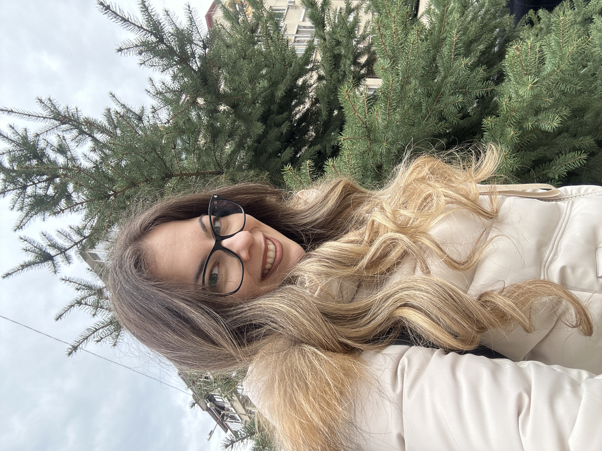 A woman poses for a selfie outdoors. She is wearing glasses and a cream-colored jacket with a fur-lined hood. Her long, wavy hair is a mix of blonde and brown. Behind her are two evergreen trees, and in the background, part of a building is visible, indicating an urban setting. Based on the location of the image, it can be pinpointed to be in the city of Stara Zagora, Bulgaria.