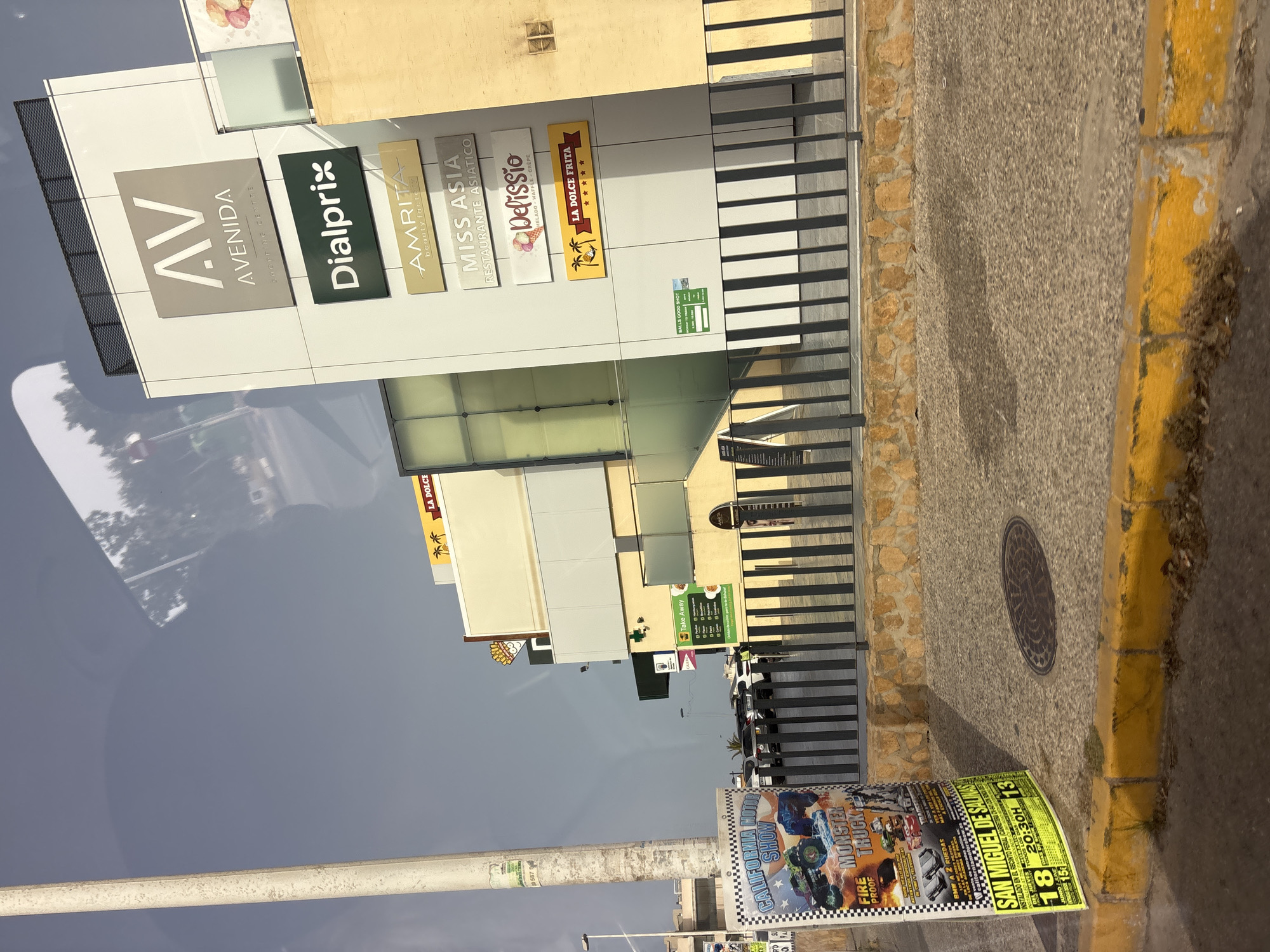 The image shows a shopping center named "AV AVENIDA SHOPPING CENTRE" located in Urbanización Villa Martín, Spain. The center has several businesses advertised on its facade, including Dialprix, Amrita beauty factory, Miss Asia RESTAURANTE ASIATICO, Delissio HELADO-WAFFLE-CREPE, and La Dolce Frita. There is also a "Take Away" sign with a menu. In the foreground, a yellow curb separates the road from the sidewalk, which features a manhole cover. A poster advertising a "CALIFORNIA MOTOR SHOW MONSTER TRUCK" event is also visible. The sky is overcast.
