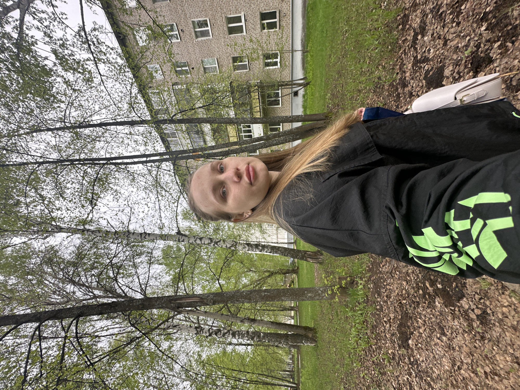 A young, fair-skinned woman with long blonde hair poses for a selfie outdoors in the city of Chaikovsky, Russia. She wears a black jacket with a bright green graphic design on the sleeve. The background includes a multi-story brick building with numerous windows, likely an apartment complex, partially obscured by bare trees with fresh green leaves. A light-colored purse is visible near the top right corner of the frame. The ground is covered with a mix of green grass and fallen brown leaves. The overall impression is a casual snapshot taken in a park-like setting within a residential area.