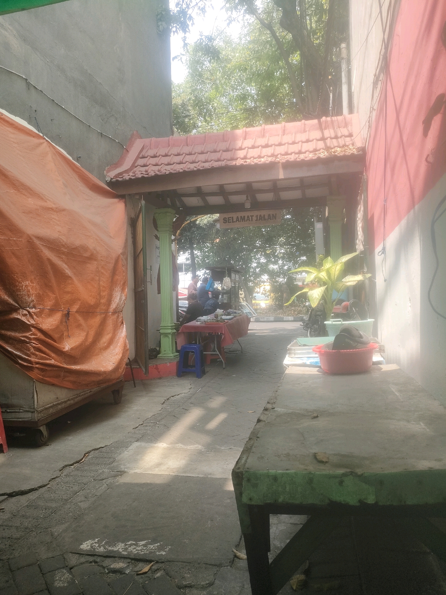 The image captures an outdoor scene, likely a street-level market or food stall area, situated in the city of Surabaya, Indonesia, based on the signage "SELAMAT JALAN" which is Indonesian for "Have a good journey" or "Goodbye". The environment appears somewhat weathered, with cracked pavement in the foreground and a large, orange tarpaulin covering a structure on the left side of the frame.
In the background, under a tiled overhang supported by green pillars, a small gathering of people is visible. They appear to be interacting around tables set up for business. One table is covered with a red cloth, and a blue plastic stool is placed nearby. There are food items and containers on the table. Further back, a yellow vehicle, possibly a bus or truck, is visible through the trees.
To the right of the frame, a red-painted wall with a white stripe runs along the building. A concrete table with a green painted edge is positioned in the foreground, and on it rests a red basin containing what appears to be a black object, possibly shoes, and a white bowl. A potted plant with large green leaves stands next to the red basin.
The overall impression is that of a modest, functional commercial space in an urban setting, possibly a rest stop or a small marketplace, with the signage suggesting a point of departure or arrival. The lighting suggests it is daytime, with sunlight casting shadows.