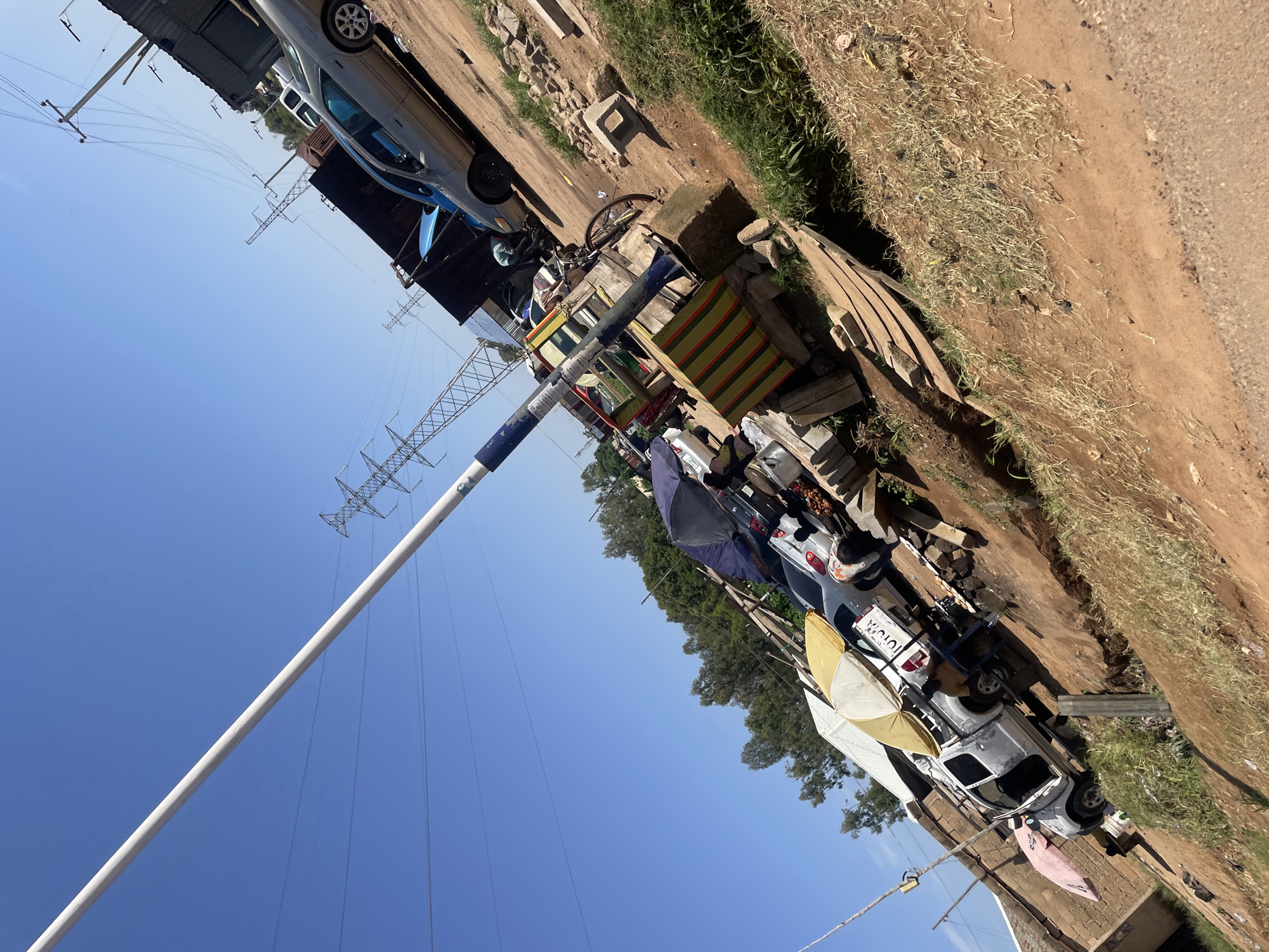 In the city of Gura Top, Nigeria, an outdoor scene depicts a collection of vehicles and market stalls under a clear blue sky. A white Toyota pickup truck, with its doors open, is parked on a dirt lot. A yellow umbrella is placed over the truck's bed. To the right of the pickup, a gray sedan is visible, along with other cars in the background. Several market stalls with colorful striped awnings are set up, some with umbrellas providing shade. A bicycle leans against a stall. In the background, power lines and pylons are prominent against the sky, suggesting an urban or semi-urban setting. The ground is a mix of dirt and dry grass.