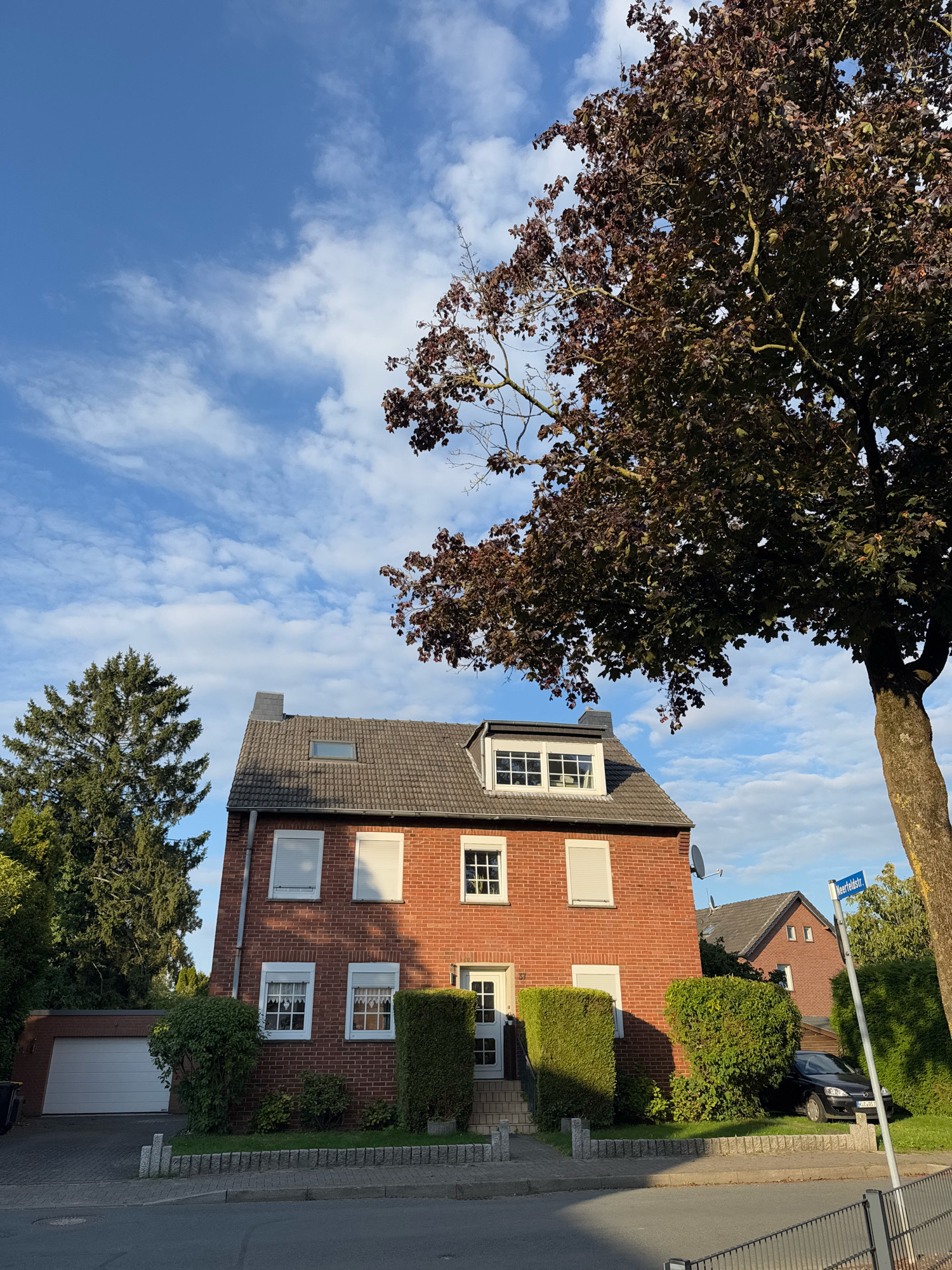 The image shows a residential street in Cleves, Germany, under a partly cloudy blue sky. A two-story brick house with a grey tiled roof is the main subject, featuring several windows with white frames. A detached garage with a white roll-up door is visible to the left of the house. A large tree with dark red leaves partially overhangs the scene from the right, and a tall evergreen tree stands to the left of the house. A street sign labeled "Meerfeldstr." is mounted on a pole on the right side of the image. A dark-colored car is parked in front of a neighboring house in the background. The scene is one of a typical quiet suburban environment.