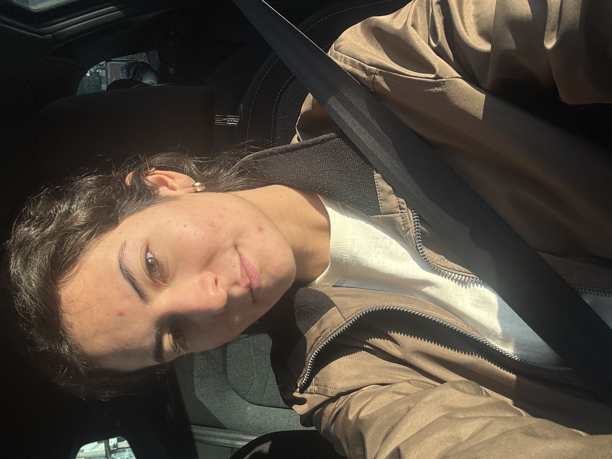 A young woman wearing a brown jacket and a white t-shirt is sitting in a car wearing a seatbelt. She is looking at the camera with a slight smile. The image was taken inside a vehicle, presumably in Puebla, Mexico, based on contextual inference. The interior of the car is visible.