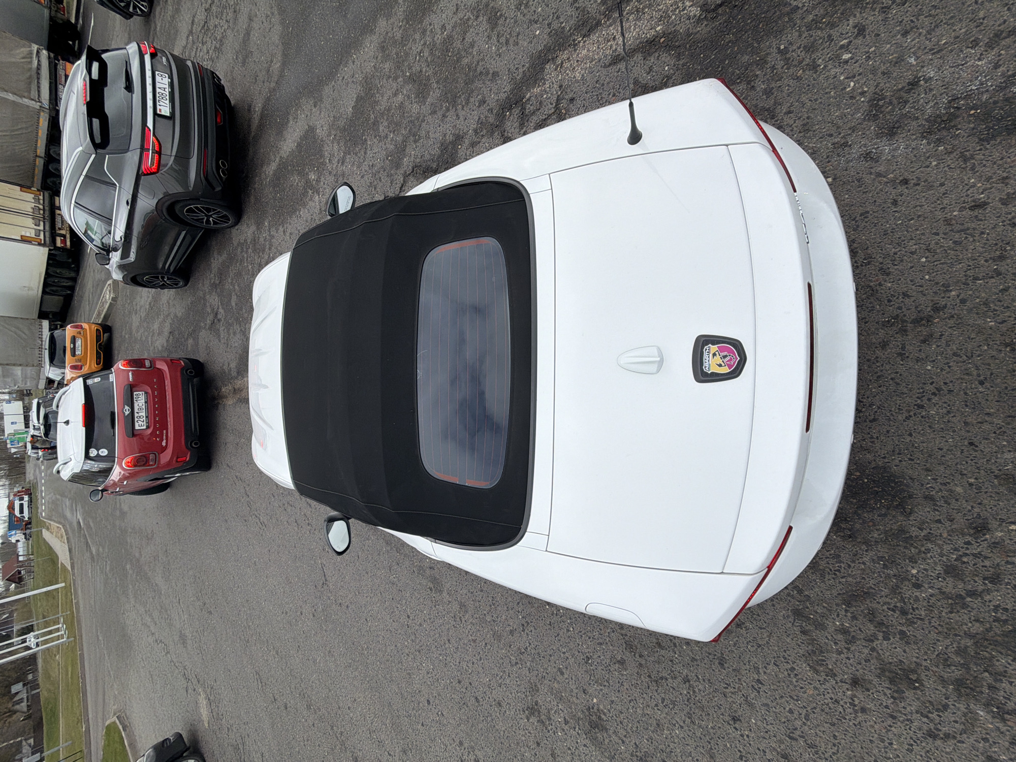 A high-angle shot shows several parked vehicles on an asphalt surface in what seems to be a parking area. The primary focus is on a white convertible with a black top, adorned with the Abarth logo on its rear. To the left, there's a red Mini Cooper and a dark grey SUV. Further in the background, there are other cars of various colors, along with large transport trucks. Given the license plates on the cars, the location is presumed to be in Дорскі сельскі Савет, Belarus. The weather appears overcast.