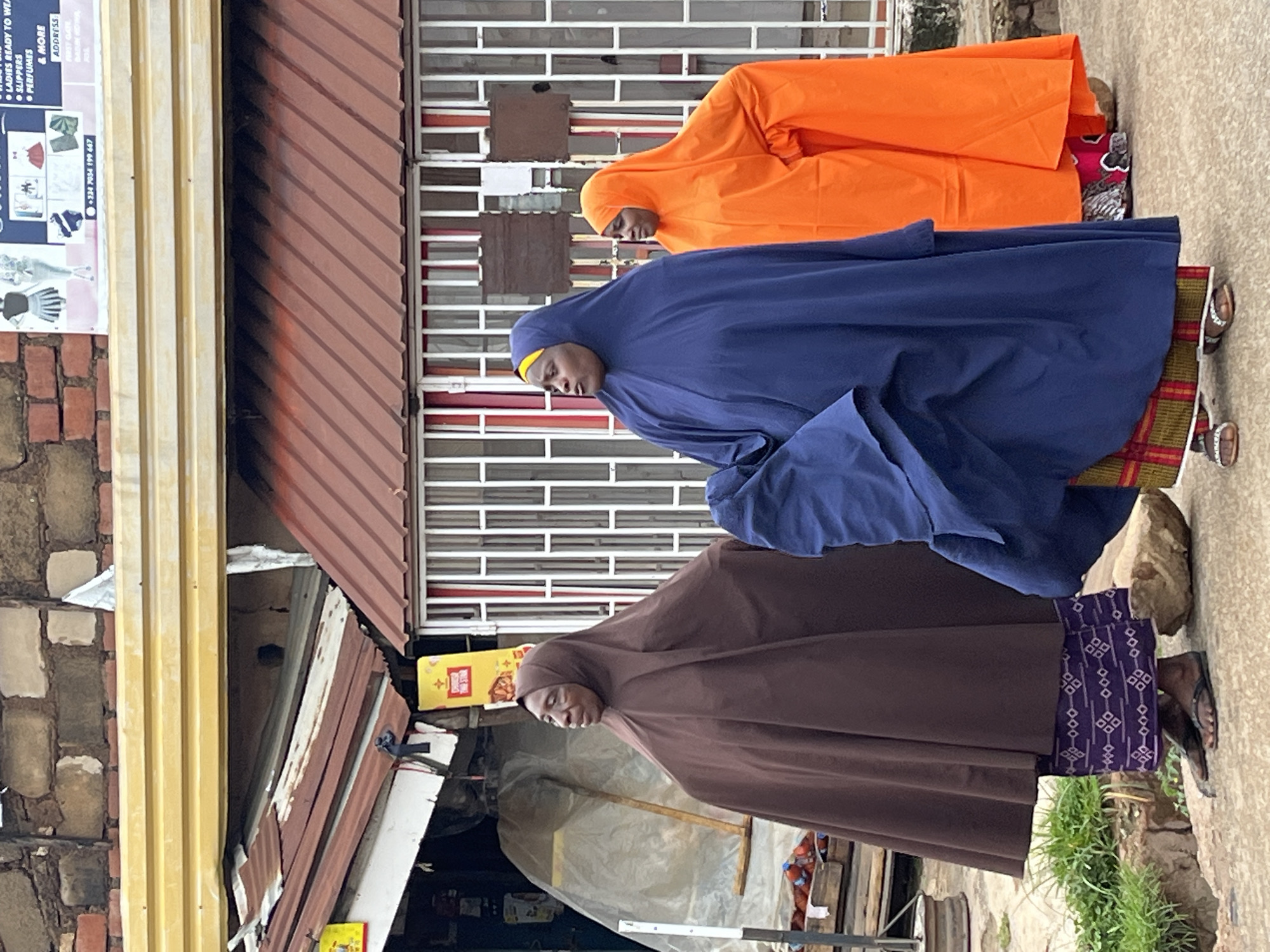 Three women, dressed in traditional Islamic attire, are standing on a paved surface in front of a building with a corrugated metal roof. The woman on the left wears a brown hijab and a patterned skirt, while the woman in the middle wears a blue hijab and a red and green patterned skirt. The woman on the right is dressed in an orange hijab. The building appears to be a shop, with metal bars on the windows. In the background, there is a brick wall and a yellow awning. A sign with text and images is visible above the awning, which indicates this location is in Jos, Nigeria, as it displays a phone number with the Nigerian country code +234 and mentions "ADDRESS FIRST GATE". The overall scene suggests a street-level view in an urban environment.