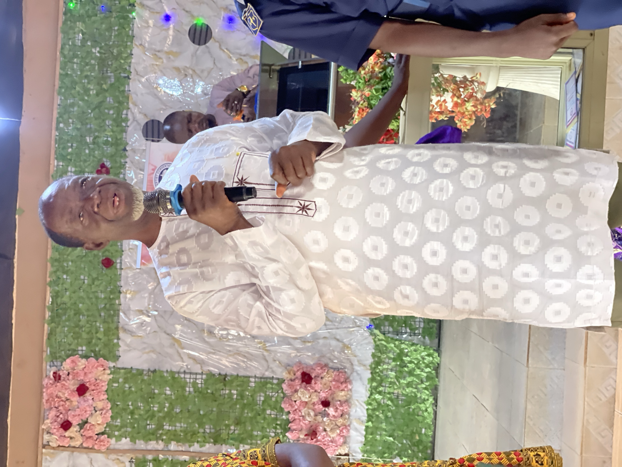 The image, rotated 90 degrees clockwise, captures an indoor event featuring a light-skinned man with a greyish beard and short hair, speaking animatedly into a microphone. He is dressed in a white, loose-fitting traditional garment, likely an agbada, adorned with subtle patterned embroidery. His mouth is slightly open, suggesting he is mid-sentence, speaking, or singing, and he appears to be in good spirits.
The background is elaborately decorated, indicating a formal gathering or celebration. It features sections of green artificial foliage panels, interspersed with clusters of pink and red artificial flowers, and shimmering white and gold patterned fabric. Small decorative lights, emitting blue and green hues, are visible at the top of the backdrop.
Several other individuals are partially visible. In the mid-ground, the head of another person is seen. Towards the top of the frame, a person wearing a blue uniform is present, while in the lower foreground, an arm with a colorful, patterned sleeve is visible, hinting at other attendees or participants. A window is seen to the right of the main speaker. A small, partially obscured sign in the background shows the text "CONVENTION" and possibly "BAPTIST," suggesting the event is a religious or organizational convention. The indoor setting and artificial lighting imply the event is taking place in the evening or night. The attire and general atmosphere are consistent with a cultural or religious gathering in Akwanga, Nigeria.