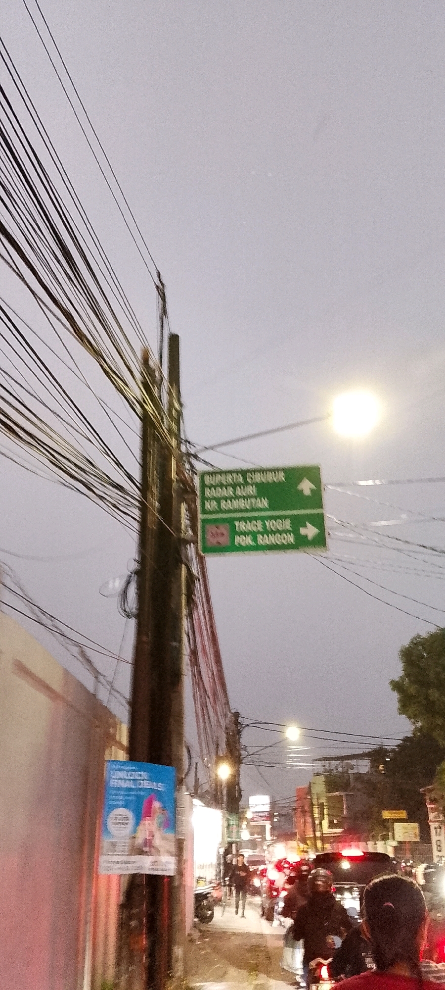 This is a medium-shot, low-angle, vertical, natural light photograph taken on a street in Depok, Indonesia, likely during twilight or an overcast day. The scene is dominated by a utility pole heavily laden with numerous black electrical and communication wires, creating a dense, tangled network that fills much of the upper portion of the frame.
Attached to the utility pole are two green directional street signs. The upper sign, with a white arrow pointing upwards, reads "BUPERTA CIBUBUR RADAR AURI KP. RAMBUTAN". The lower sign, with a white arrow pointing to the right and a small red circular emblem, reads "TRAGE YOGIE POK. RANGON".
To the left of the utility pole, a bright blue poster is affixed to it, displaying the text "UNLOCK FINAL DEALS" and an image of people with vibrant pink hair.
The street below is congested with traffic, primarily motorcycles, with some cars visible. Brake lights from the vehicles cast a red glow on the wet asphalt. Several individuals are seen on motorcycles, some wearing helmets, and others walking. The atmosphere suggests a busy urban environment, possibly experiencing a light rain or post-rain conditions due to the sheen on the road.
Buildings line the street on both sides, with storefronts and other signage visible in the background, partially obscured by the traffic and overhead wires. A street lamp is illuminated on the right side of the frame, emitting a bright, diffused light. The sky is a pale, overcast gray.