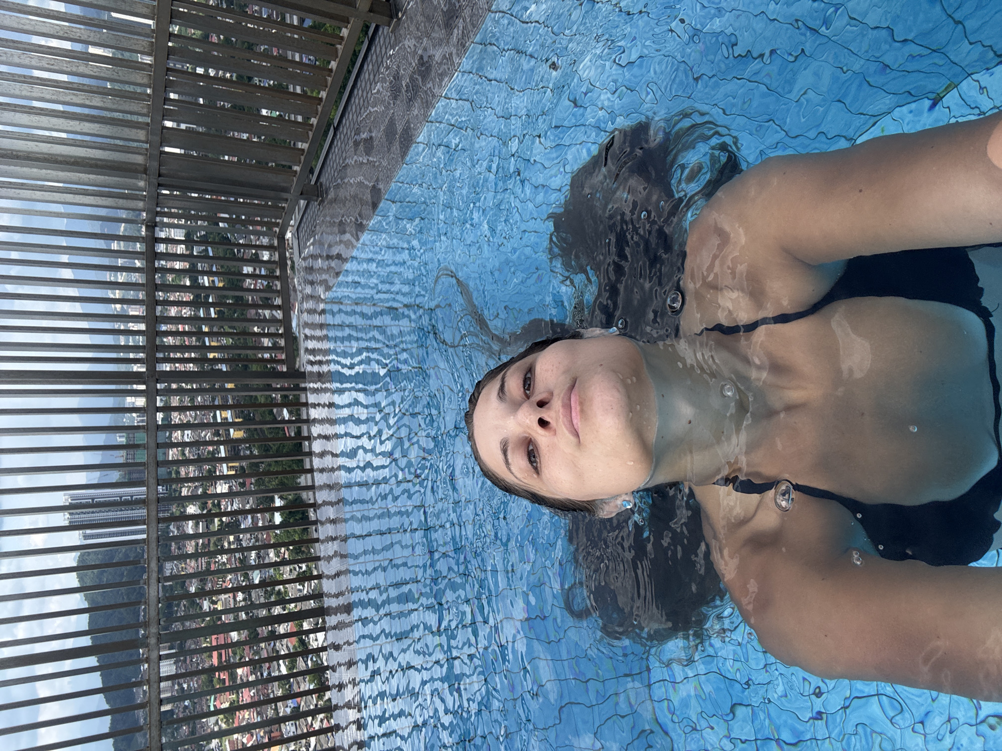 The image captures a woman floating on her back in a bright blue swimming pool, seen from a slightly elevated perspective. Her head and upper chest are visible above the water, while the rest of her body is submerged. She has long, dark hair spread out around her in the water, and her eyes are open, looking upwards with a serene expression. She wears a dark-colored swimsuit top.
The pool water is clear, revealing a light blue tiled pattern beneath the surface and showing ripples and small air bubbles around the woman's face and arms. To the left, a slatted metallic railing or fence provides a barrier to an expansive vista. Beyond this railing, a vast urban landscape unfolds, featuring numerous buildings and pockets of greenery, suggesting a densely populated area. Distant mountains rise against a clear, bright sky. The scene is bathed in natural daylight, indicating daytime. The elevated viewpoint strongly suggests a rooftop or high-floor pool, possibly an infinity pool, overlooking the city of Ampang Jaya, Malaysia. There is no visible text in the image. The overall impression is one of relaxation and calm in a scenic setting.