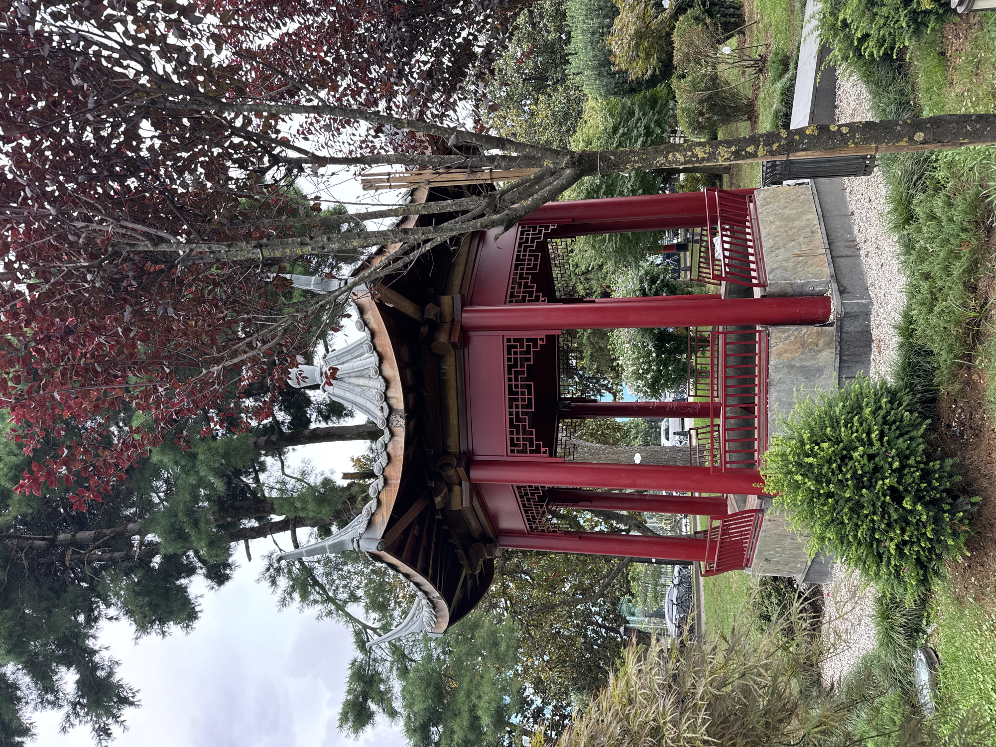 The image shows a red, octagonal gazebo with a tiled roof, located in a park in Batumi, Georgia. The gazebo has ornate detailing on its roof and lattice-work on its walls. It is surrounded by lush greenery, including trees with red and green leaves, bushes, and grass. In the background, there are cars and bicycles parked along a street. The sky is overcast. The gazebo appears to be a decorative structure within a public park, possibly a Chinese-style garden, given its architectural design.