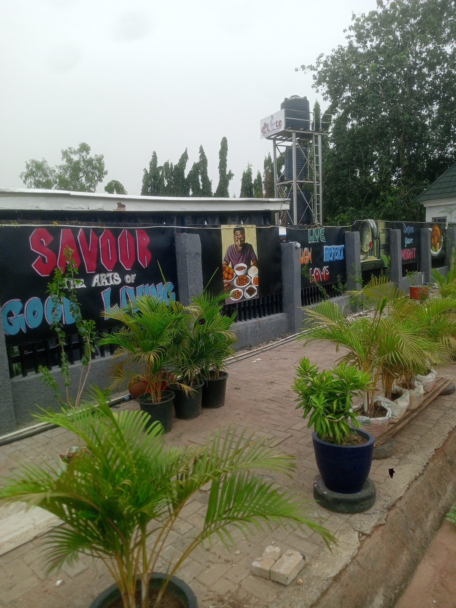 The image shows the exterior of a building in Gwarinpa, Nigeria. A painted black wall with grey pillars runs along the length of the shot, with the words "SAVOOR - THE ARTS OF GOOD LIVING" and other phrases. Illustrations of food and people are interspersed along the wall. Potted plants of various sizes line the wall and edge of a brick-paved pathway. In the background, a water tower with the word "Elite" is visible, along with trees and parts of other buildings. The sky appears overcast.
