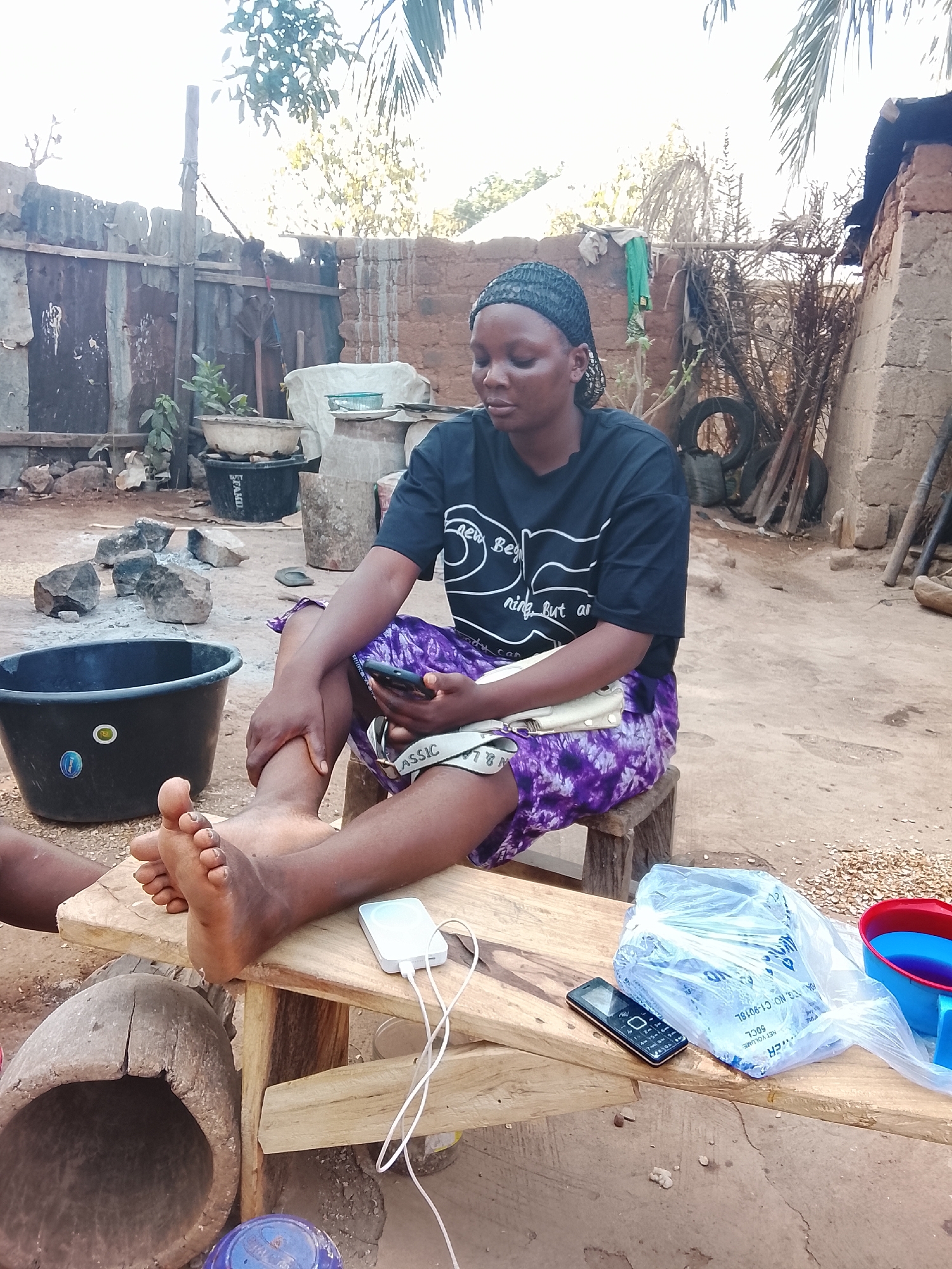 A medium shot captures a woman seated on a small wooden stool in an outdoor setting. She is dressed in a black t-shirt with white text and a design, paired with purple and white shorts. Her feet are bare, resting on a wooden bench, where a white power bank with connected cables and a basic cellular phone are also placed.
The woman is looking down at her smartphone, which she holds in her left hand, while her right hand seems to be touching her left leg, as if massaging it. A black bucket sits to the left of the stool, and a wooden mortar lies on the ground. On the right side of the bench, there's a plastic bag and a red and blue bucket.
The backdrop consists of a rustic outdoor space. There is a fence constructed with corrugated metal sheets, and a weathered brick structure stands behind it. Foliage and trees are visible in the background. The ground is a mix of soil and scattered debris, contributing to the rural, perhaps less-developed, environment.